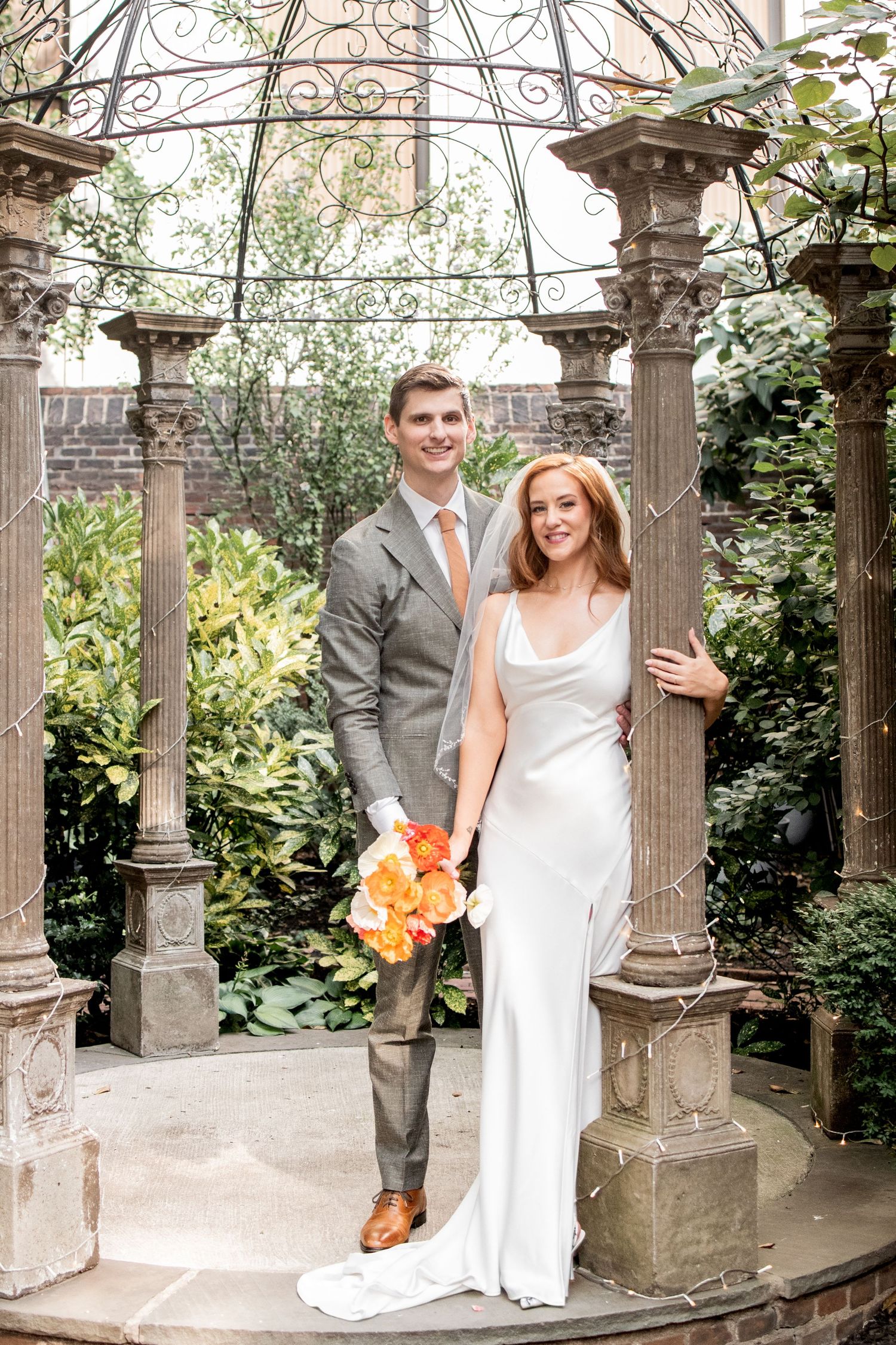 A couple celebrates their wedding day in a garden setting with ornate stone columns and lush greenery backdrop.