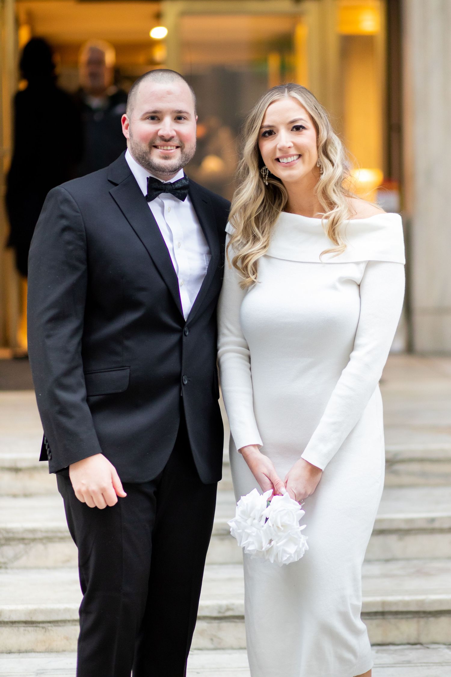 A couple poses together on stone steps, with the person wearing an elegant white long-sleeved wedding dress.
