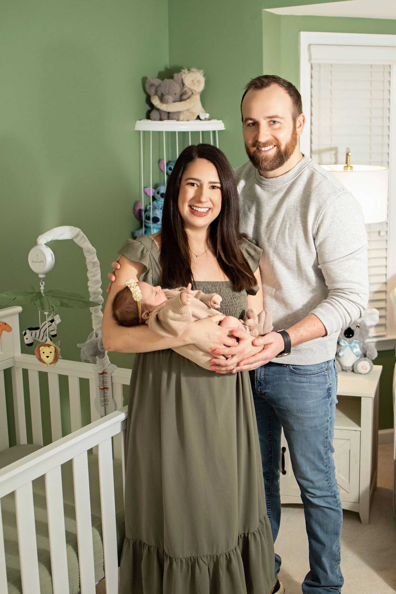 Happy couple stands in mint green nursery holding their newborn baby beside white crib.