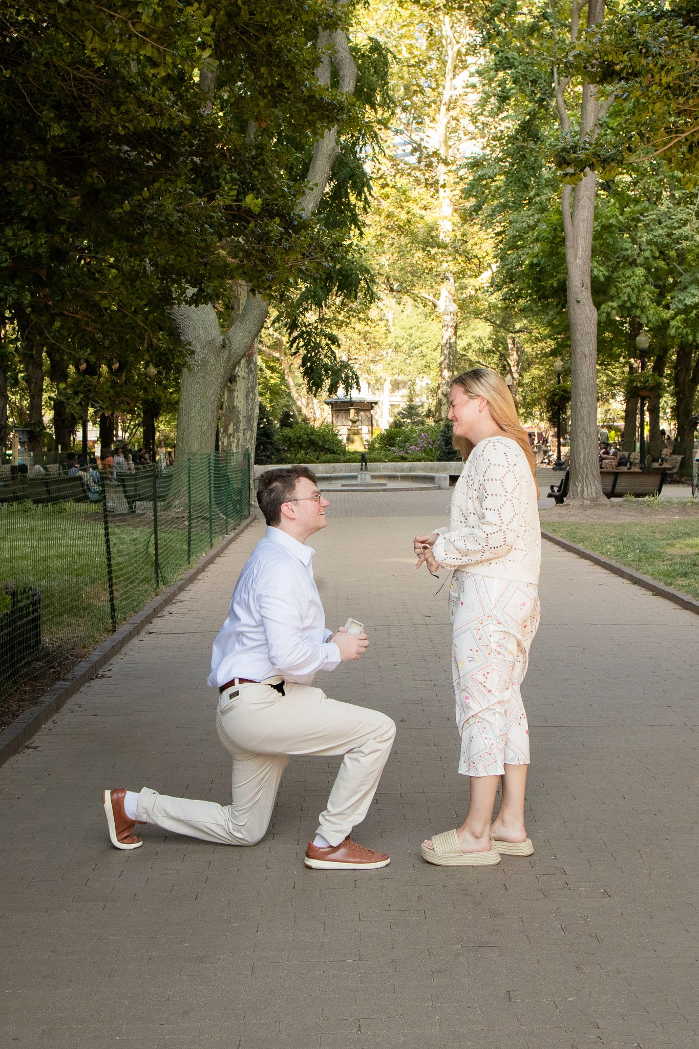 A romantic proposal scene unfolds in a tree-lined park as a person kneels before another on a paved pathway.