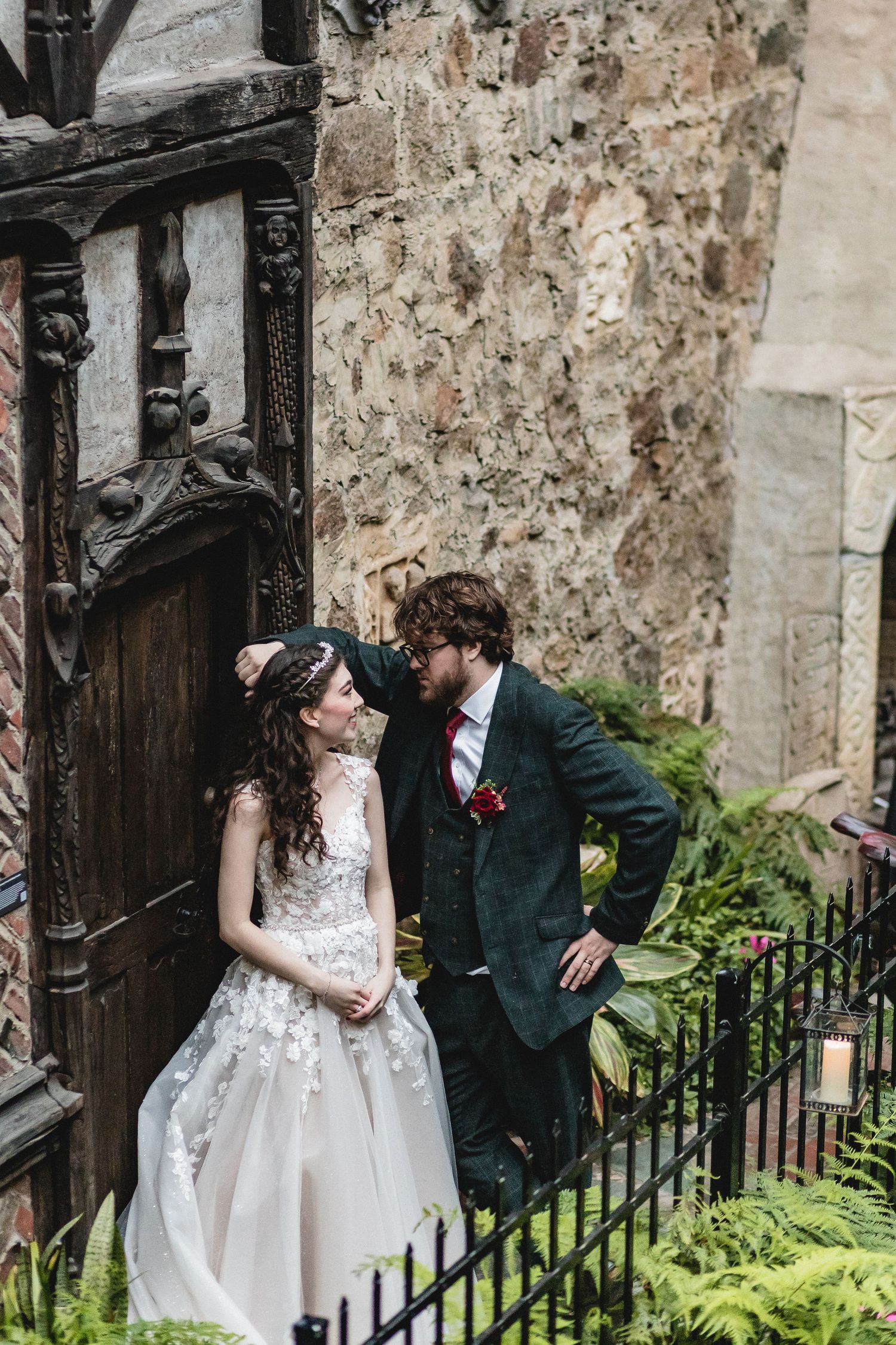 A newlywed couple stands by an old wooden door and stone wall among decorative greenery and lanterns.