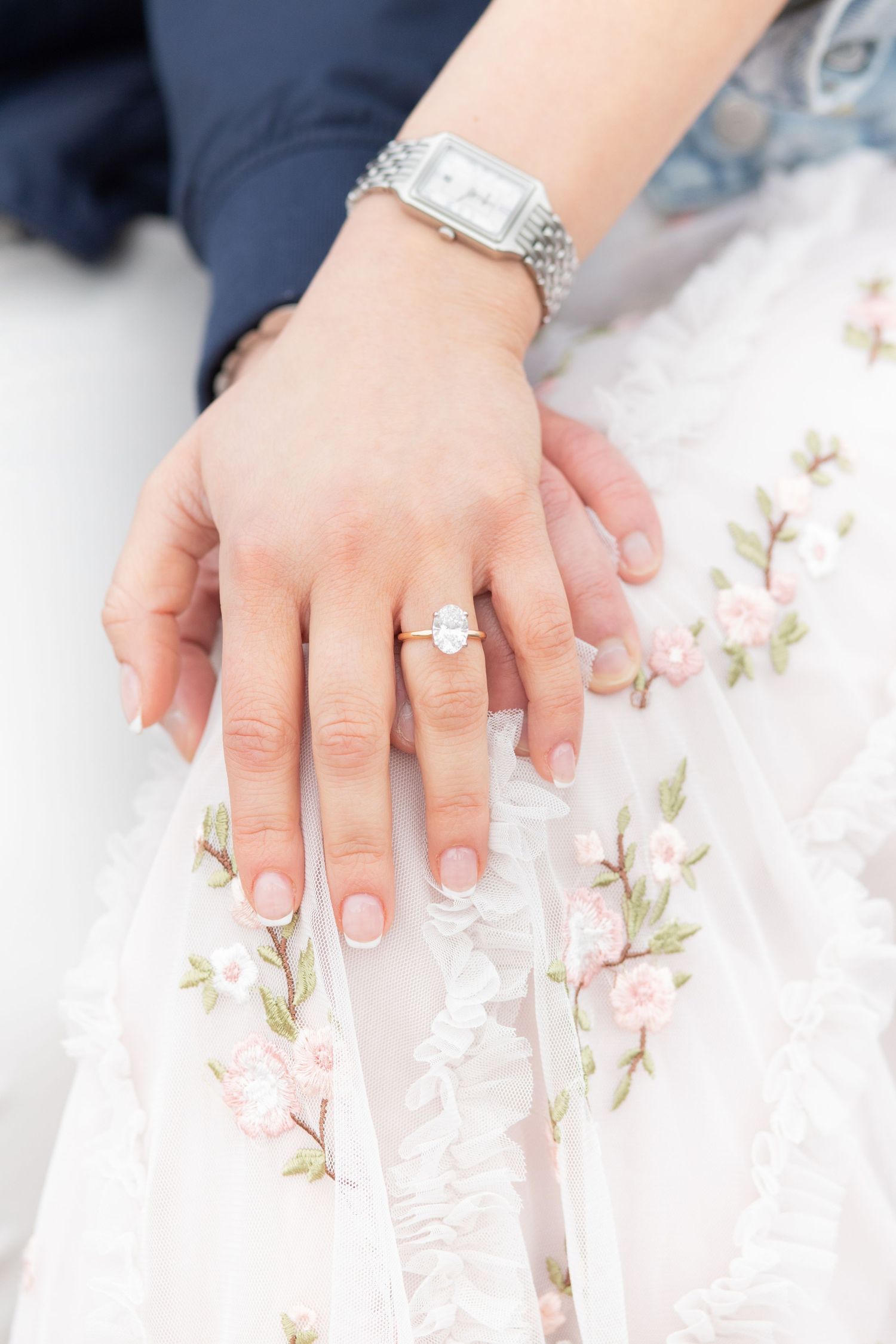 Silver watch and engagement ring detail shot on white lace dress with delicate pink floral embroidery.