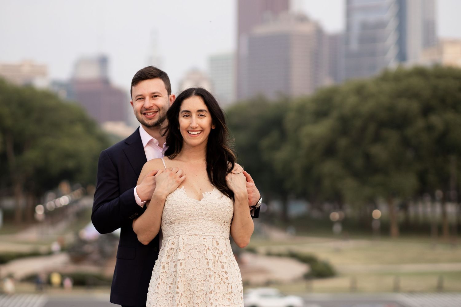 A couple poses together outdoors in front of a cityscape with greenery and buildings in the background.