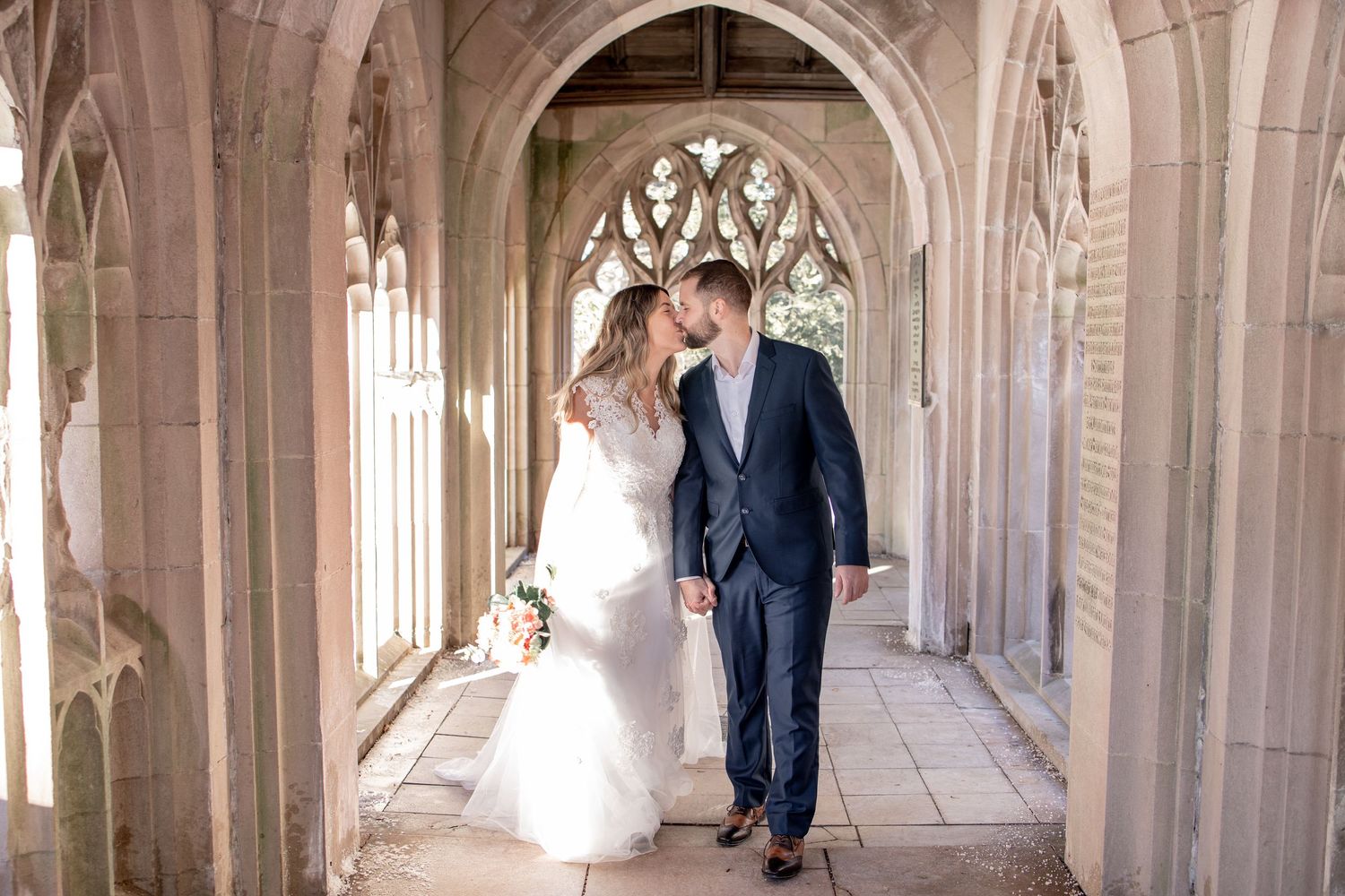 Two newlyweds walk together through a sunlit Gothic stone corridor with arched windows and vaulted ceiling.