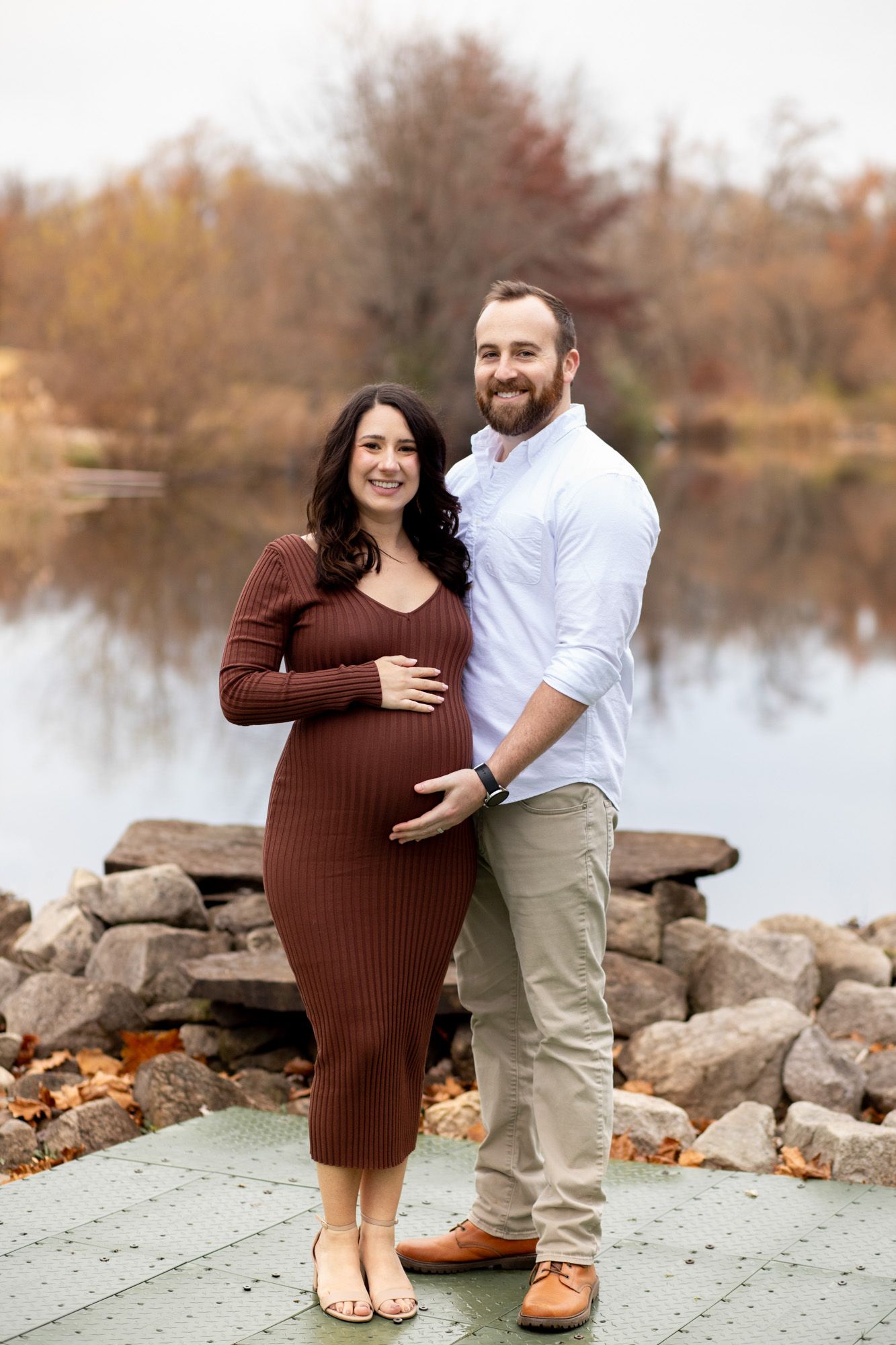 A smiling pregnant couple poses together by a lakeside with autumn foliage in the background.