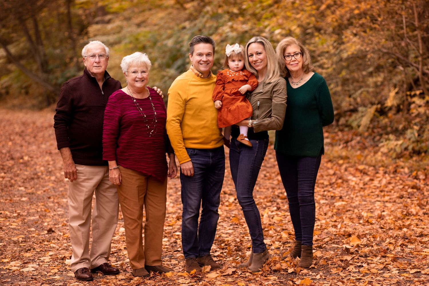 A group of people posing joyfully together on fallen autumn leaves in a wooded setting at golden hour.