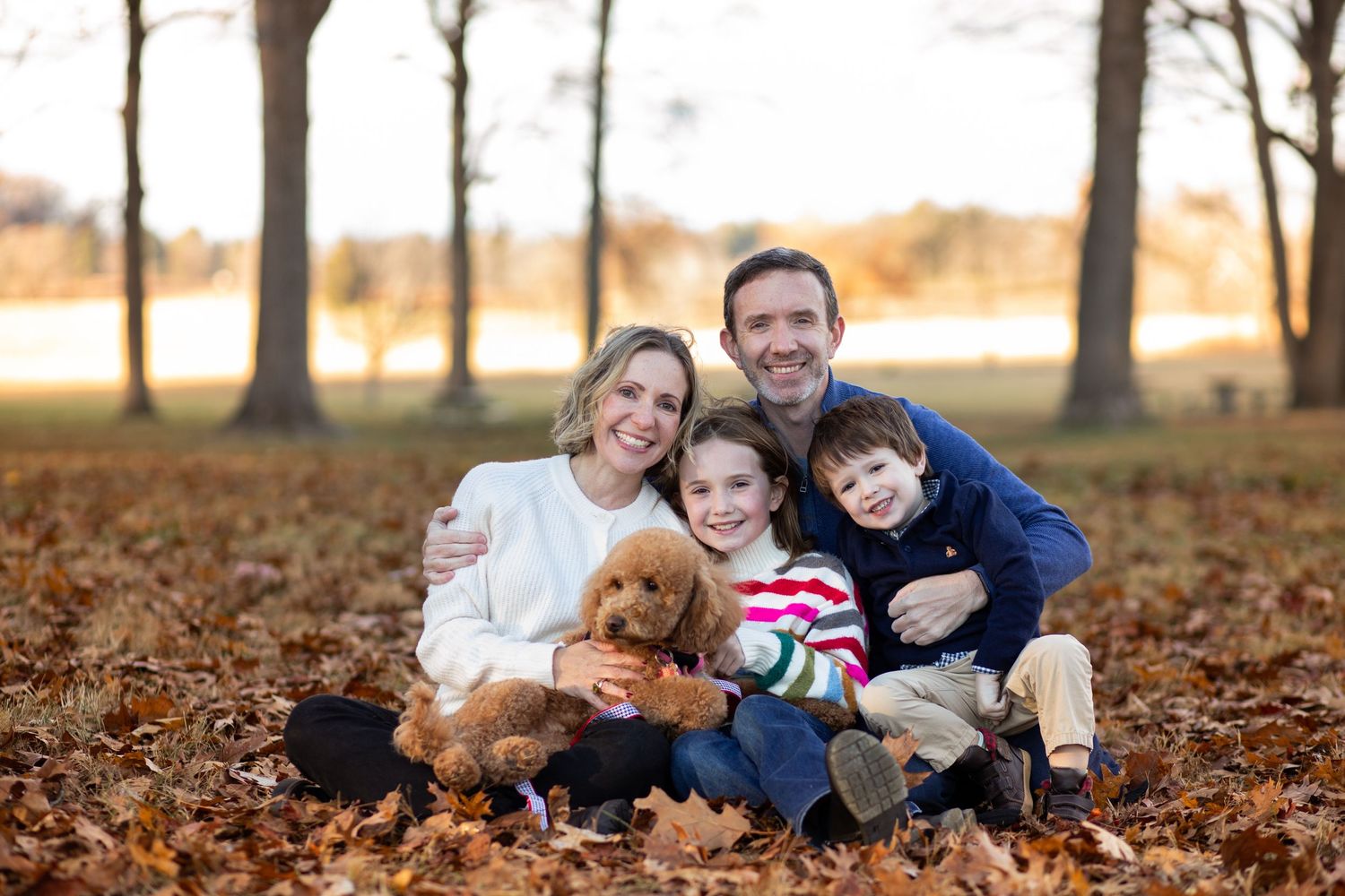 Family and golden doodle puppy sitting together on autumn leaves in a park during golden hour.