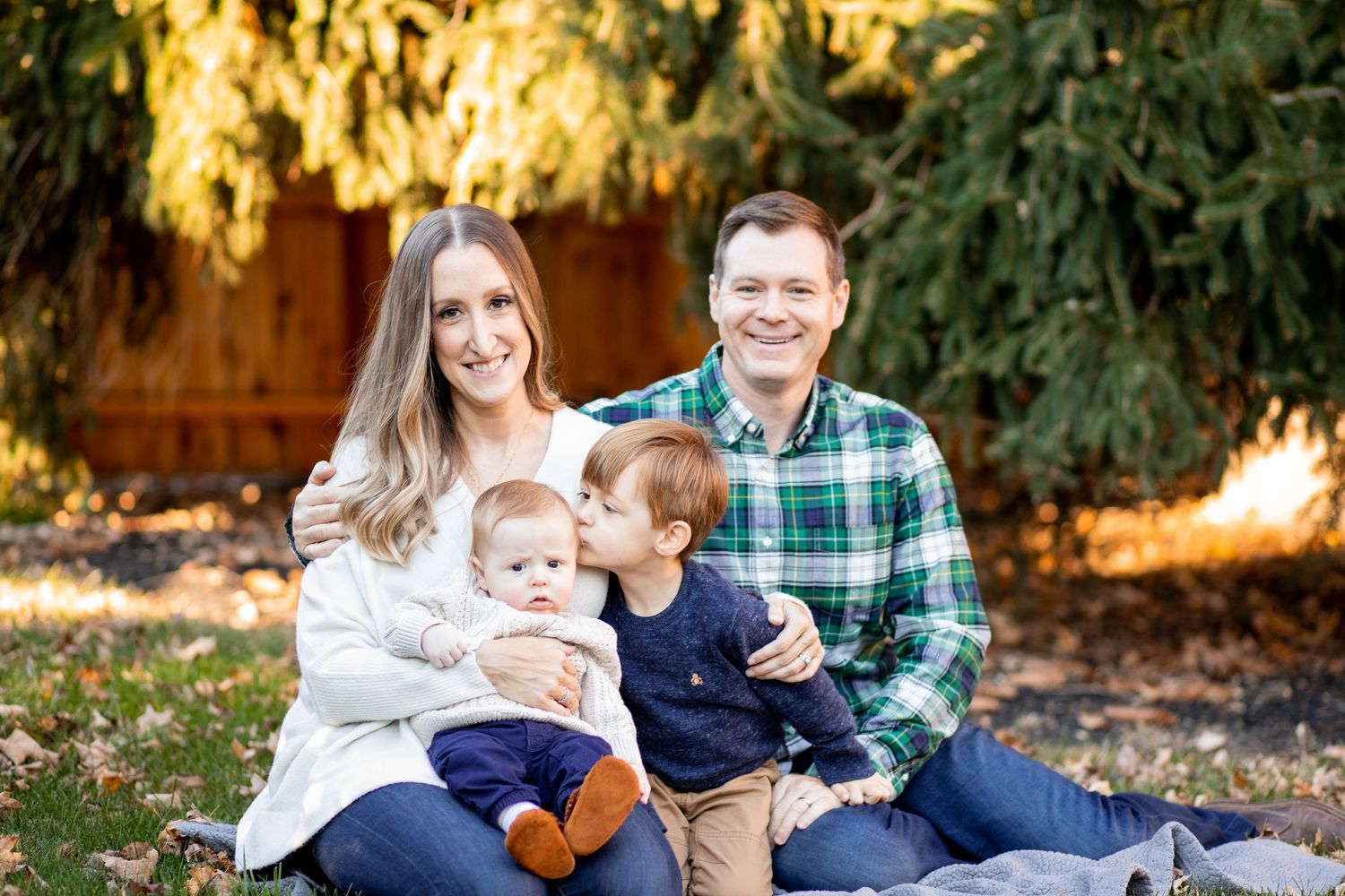 A family sits together outdoors on autumn grass surrounded by evergreen trees in late afternoon sunlight.