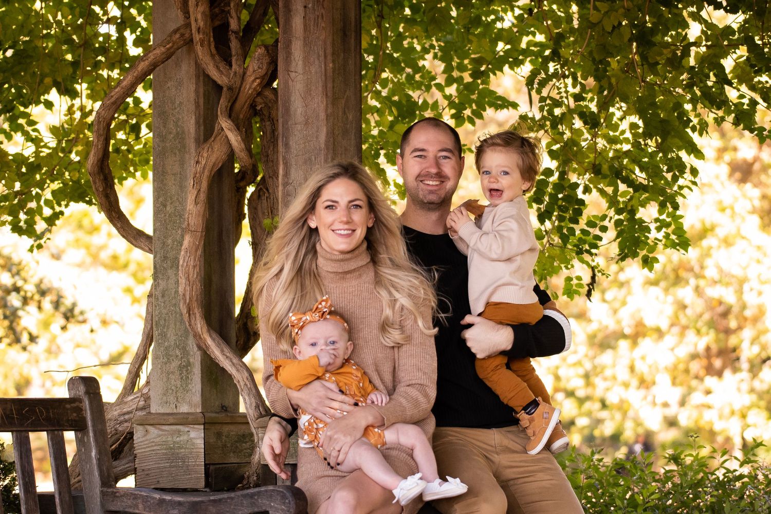 A family sits together on a wooden bench under autumn trees in warm golden light during an outdoor portrait session.