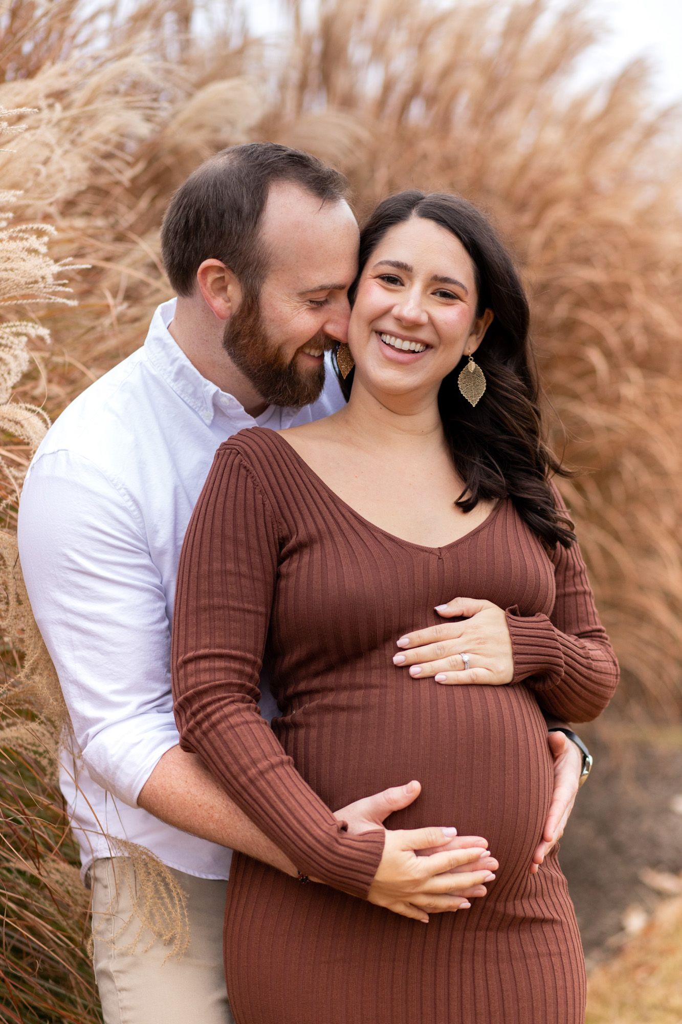 Smiling pregnant woman in brown dress embraced by bearded man outdoors among golden pampas grass during maternity photo session.