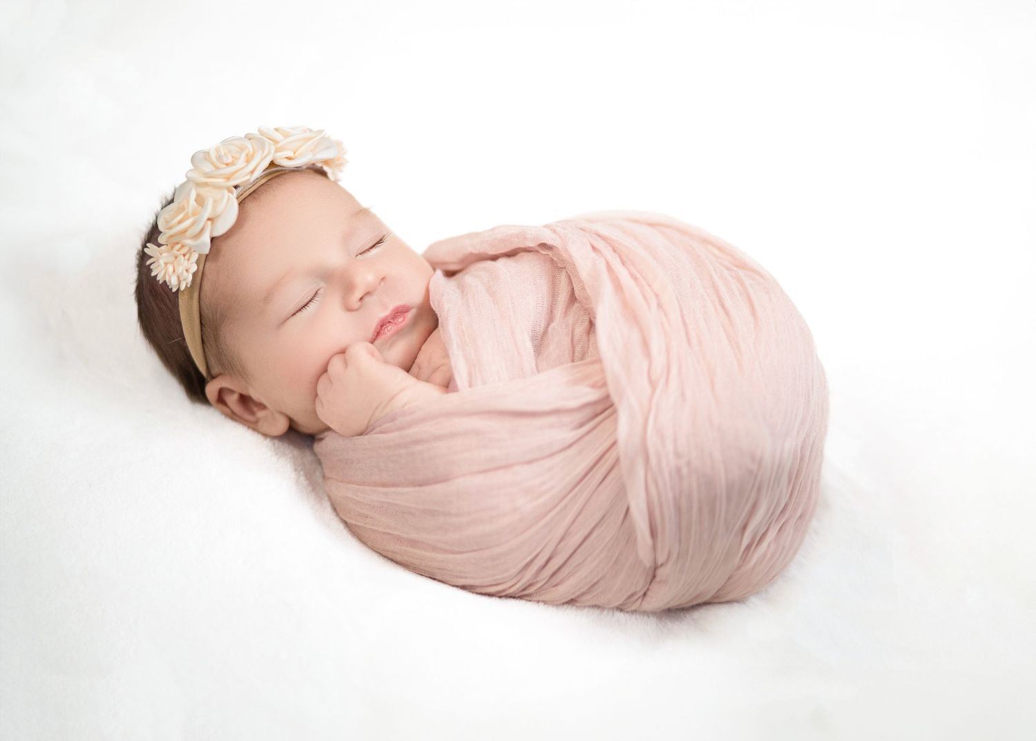 Newborn baby swaddled in soft pink fabric with lace headband sleeps peacefully on white background.