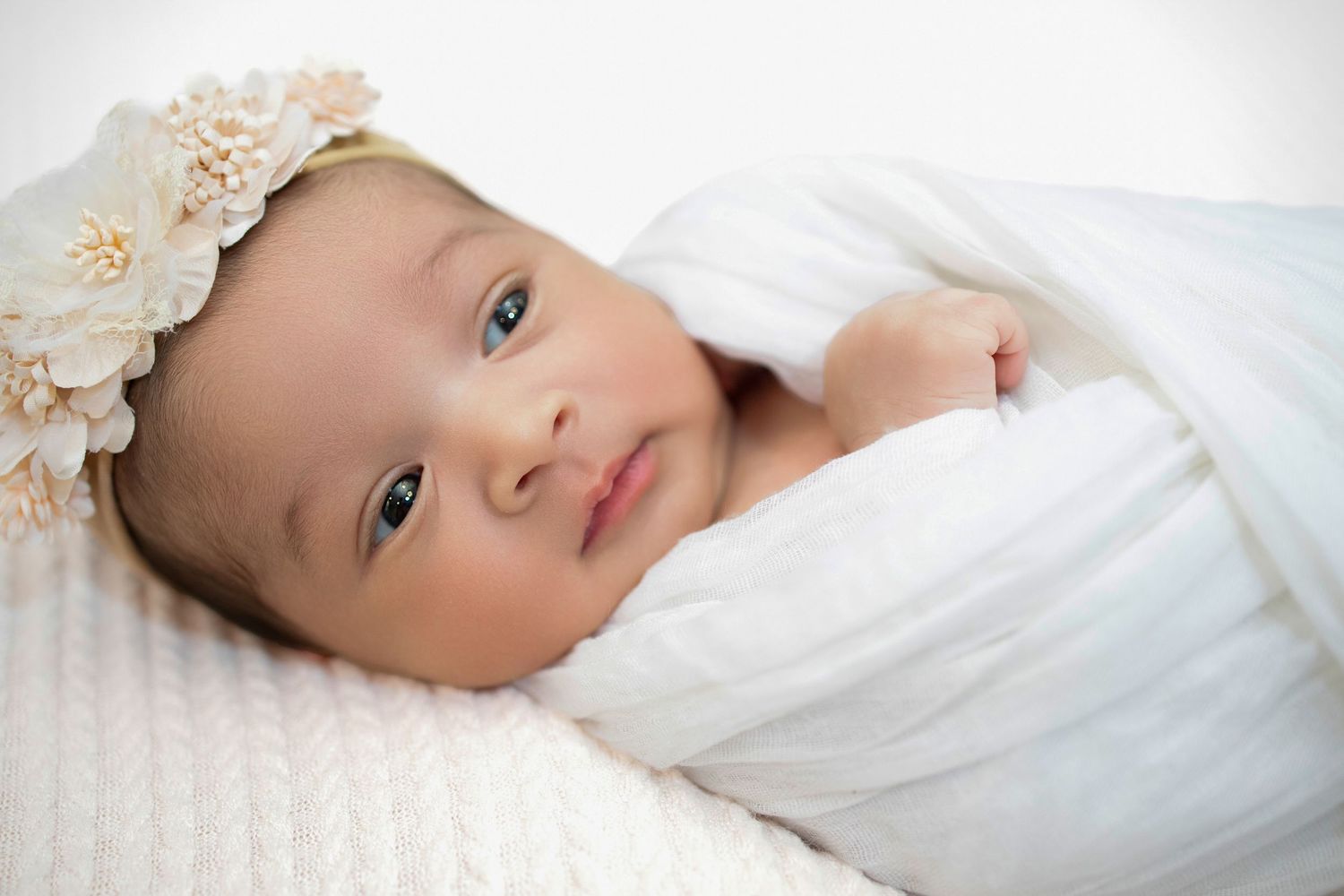 Small baby wearing a delicate lace headband lies peacefully on white blankets while looking at camera.