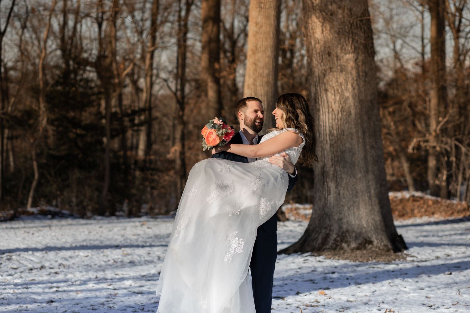 A couple in wedding attire embraces in a snowy winter forest setting with tall trees in the background.