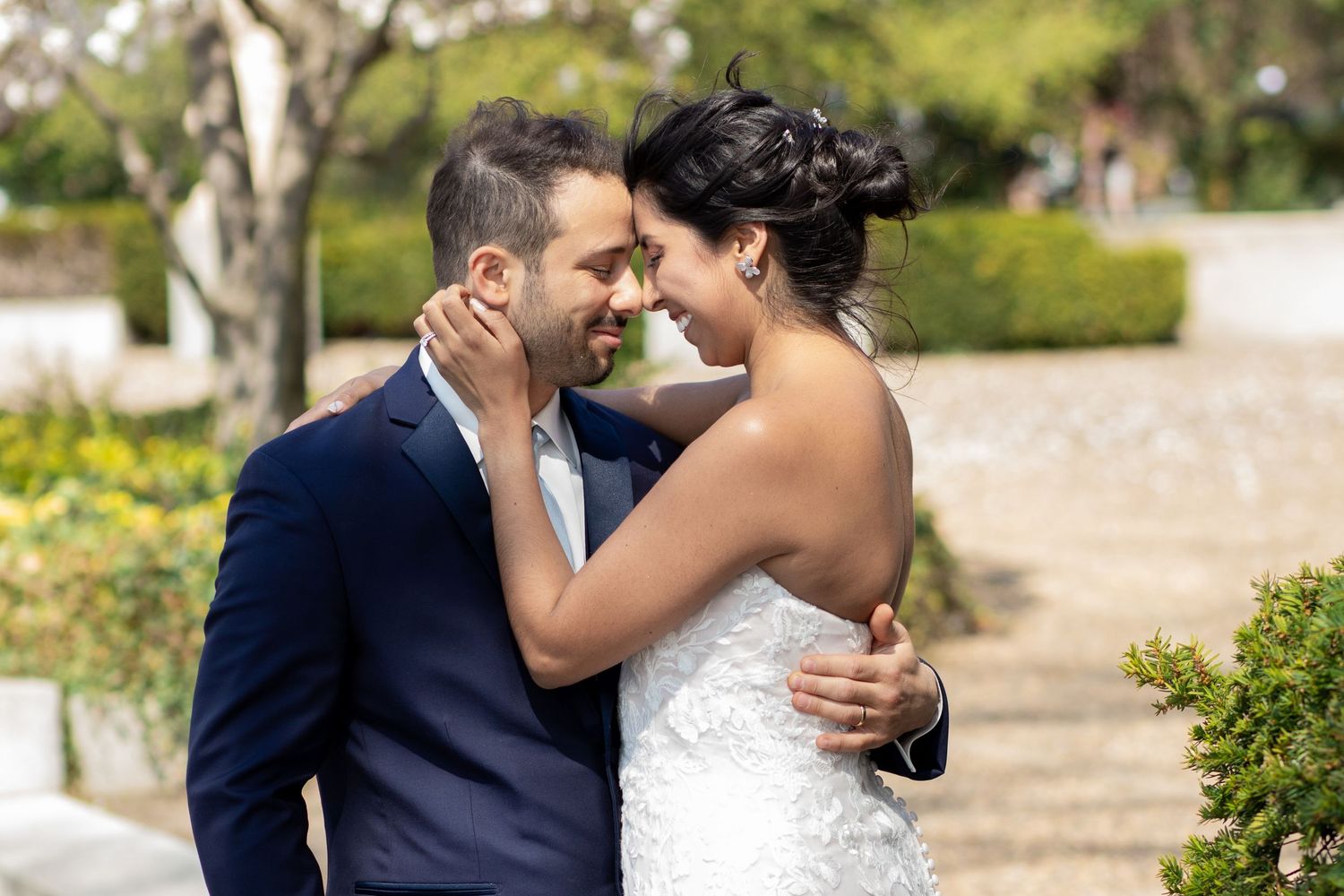 A couple shares an intimate moment in their wedding attire outdoors in a garden setting with blooming flowers.