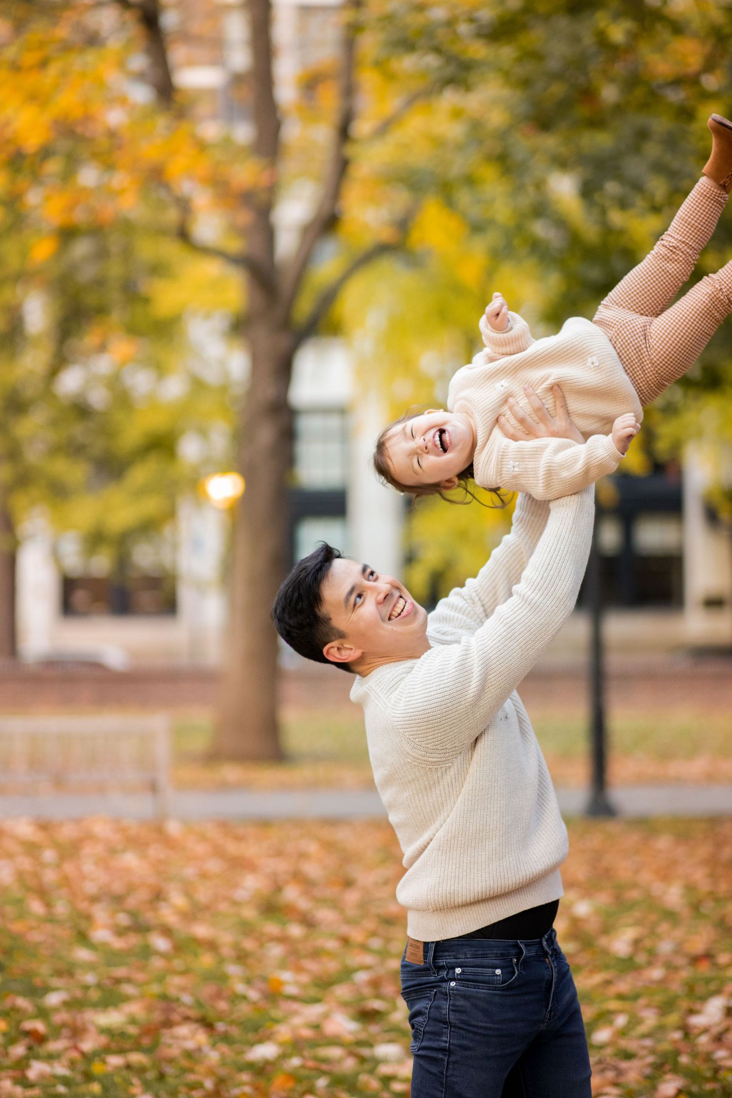 A parent lifts a young child in a cream sweater playfully in a park surrounded by autumn leaves and golden trees.