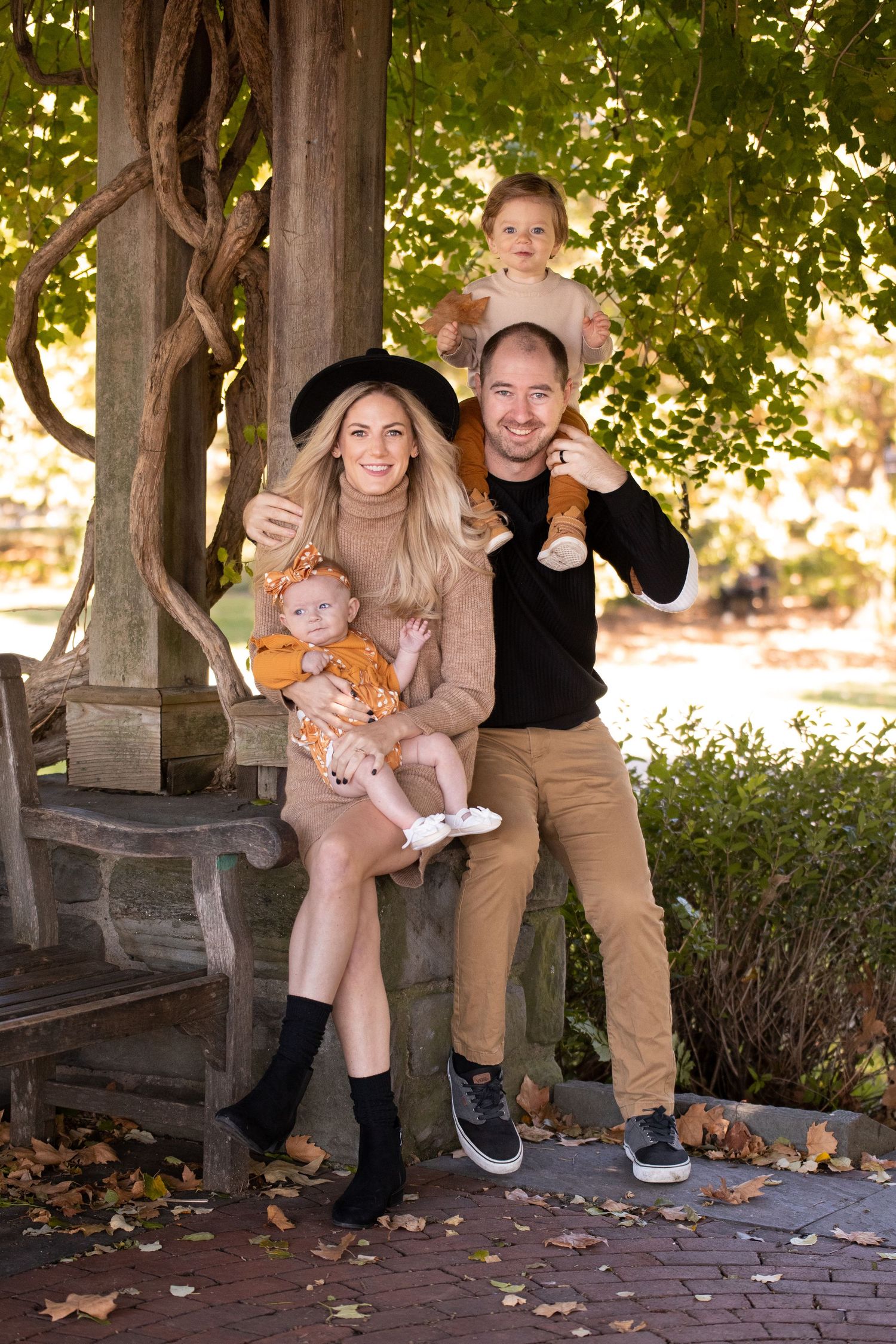 Family of four posing together at an outdoor bench in autumn with fallen leaves and greenery in background.