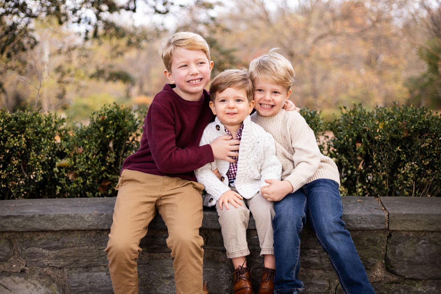Three young children sitting together on a stone wall with hedges and autumn trees in the background.