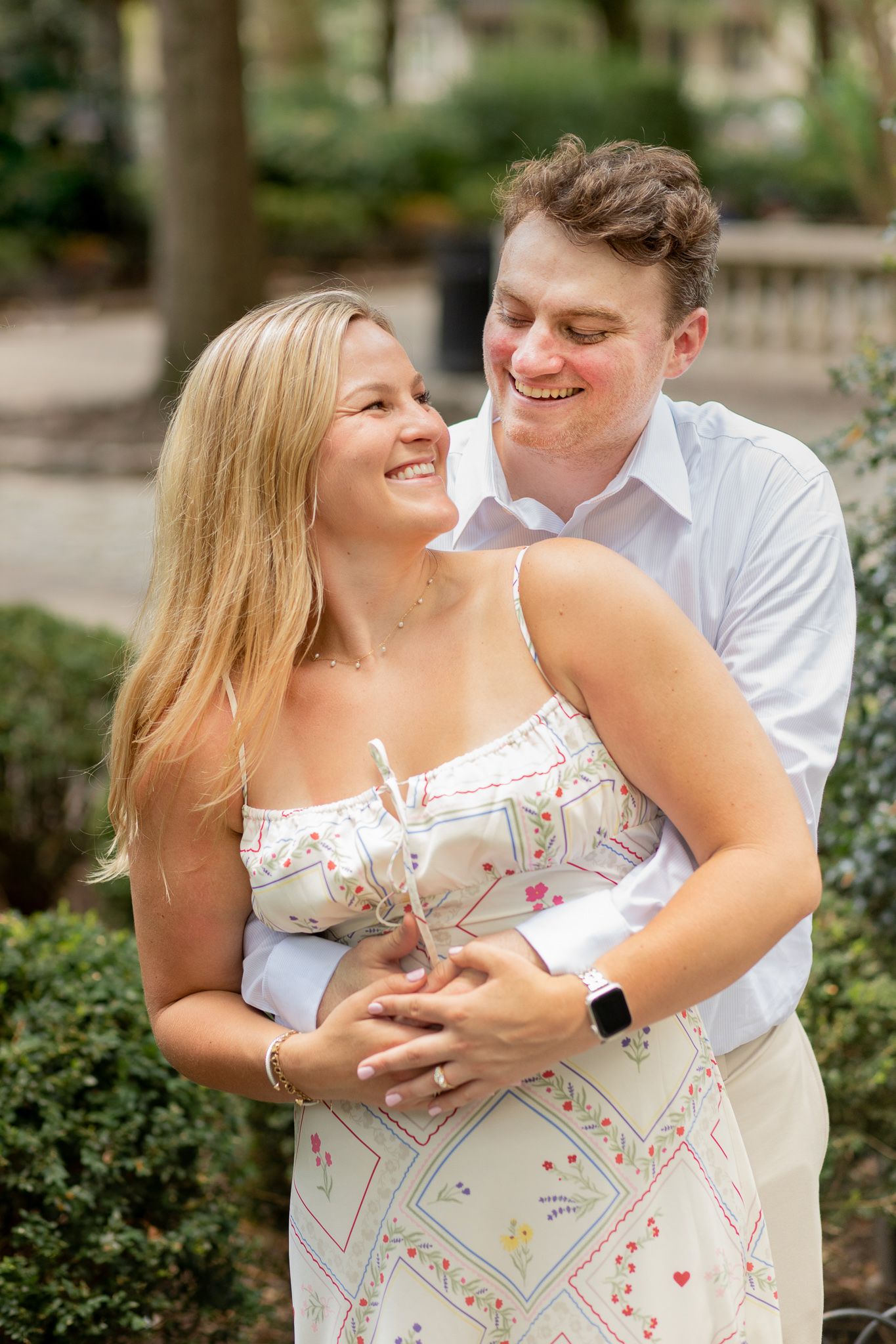 A happy couple embraces and laughs together in a garden setting with greenery in the background.