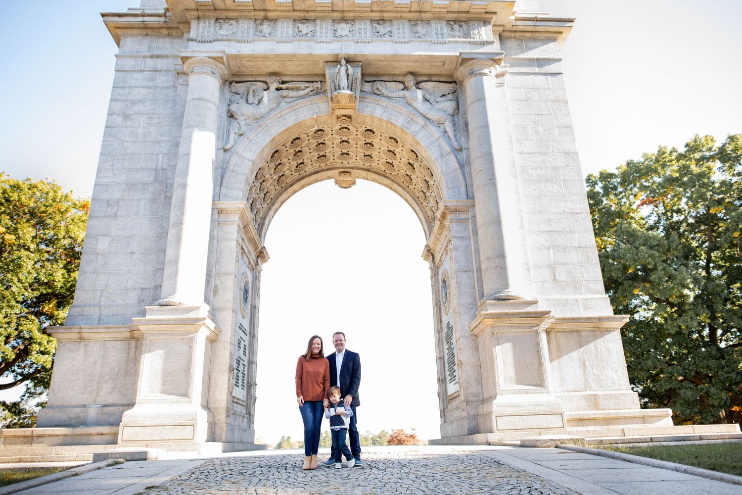 Two people stand beneath a grand marble memorial arch on a cobblestone path surrounded by greenery.
