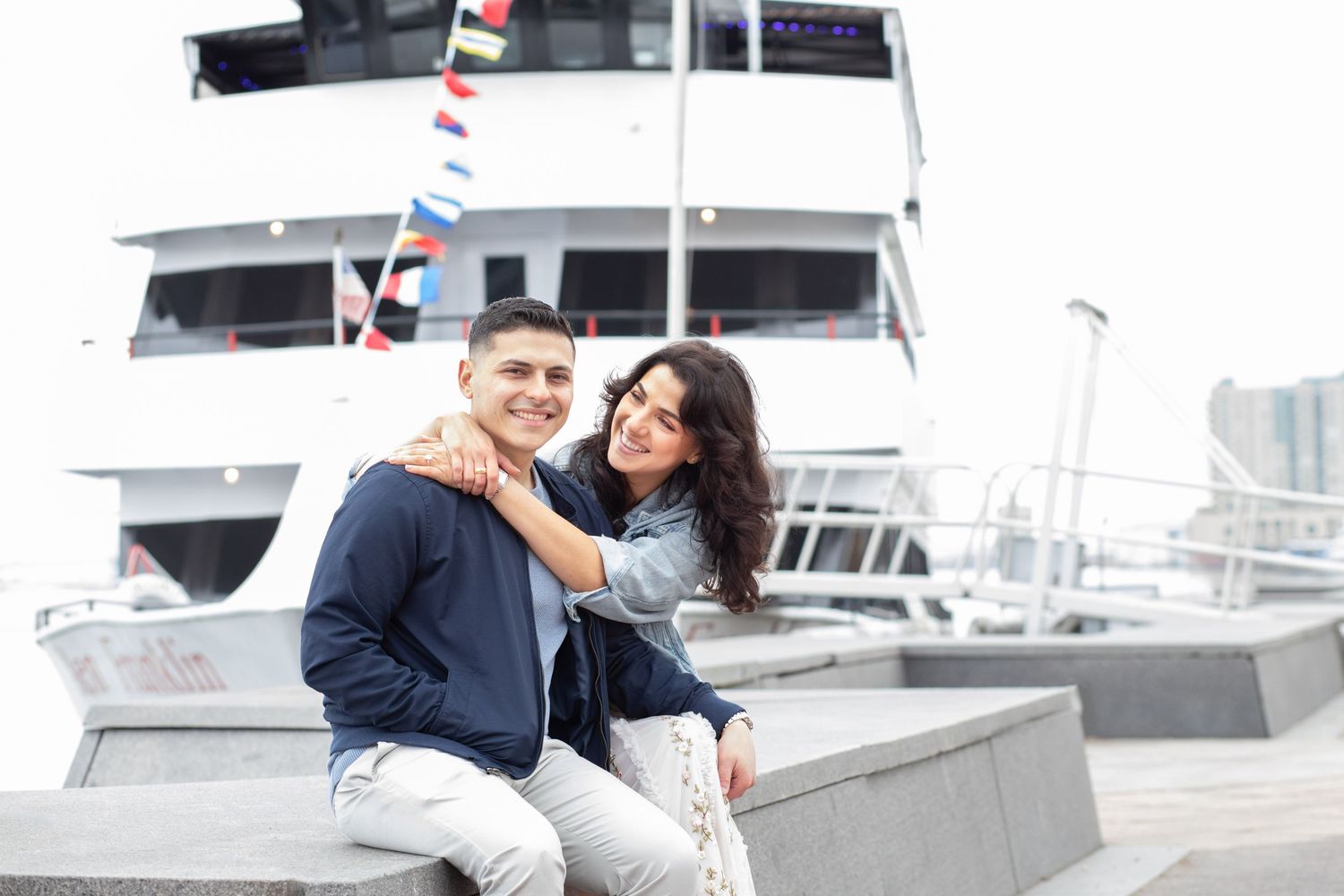 A couple sits together on a dock with boats and a marina in the background on a cloudy day.