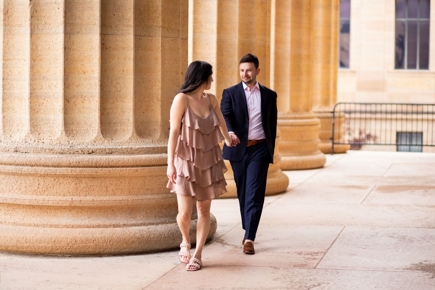A couple strolls together along a grand colonnade with beige stone columns at an elegant architectural setting.