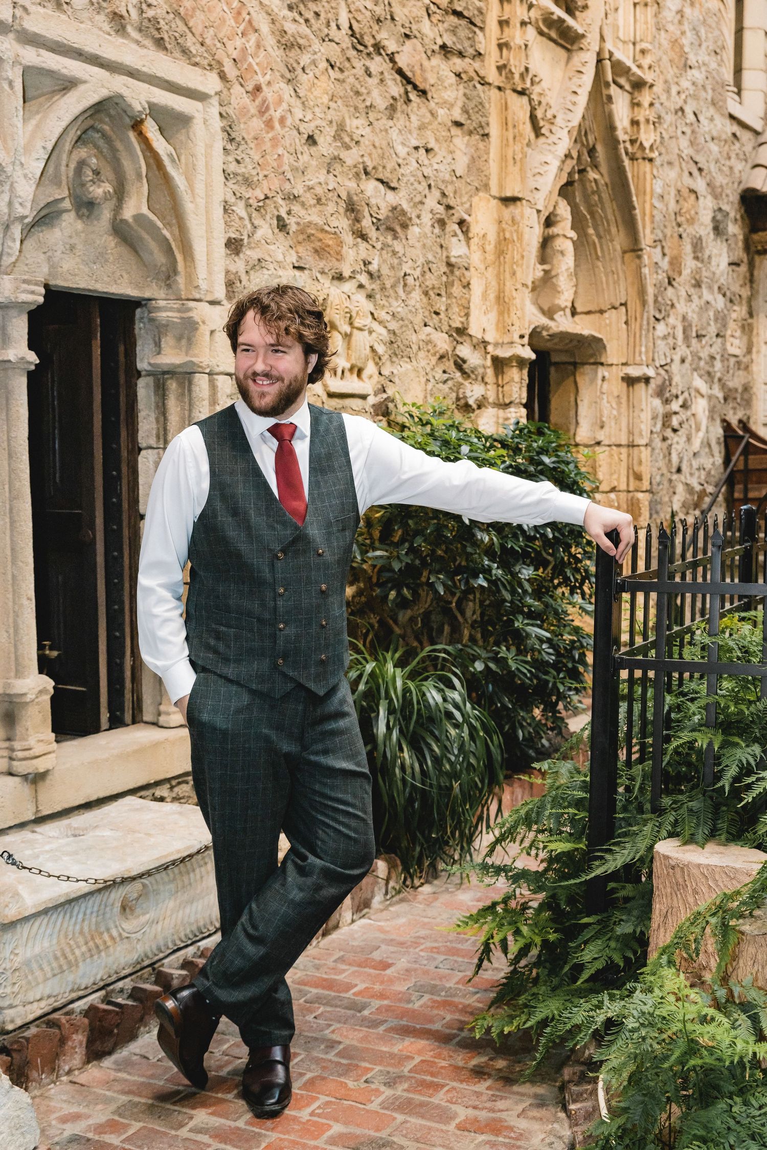 Stylishly dressed person in green plaid vest and red tie poses casually against stone church exterior with gothic arches.
