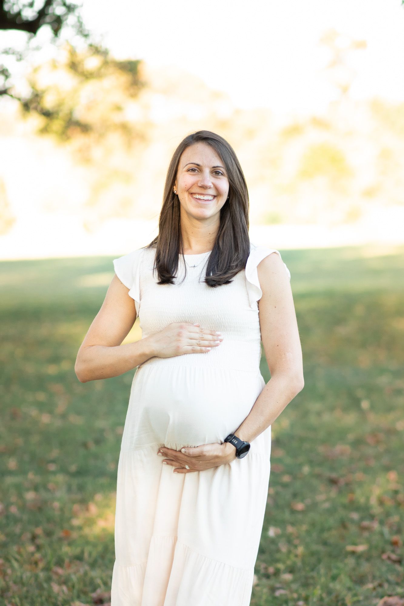 Smiling pregnant woman in white dress cradling baby bump outdoors in sunny park setting.