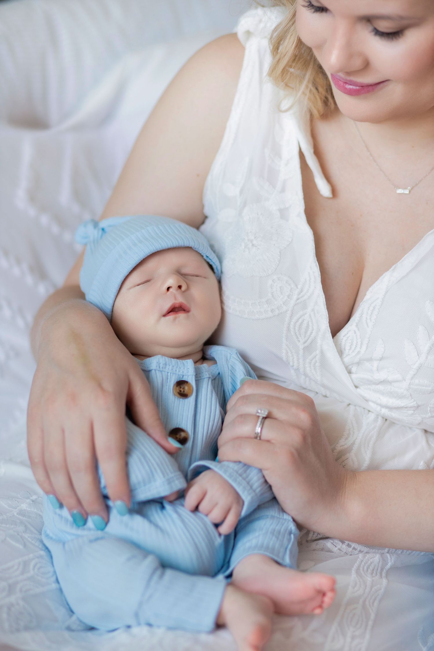A tender moment between parent and sleeping baby in white lace dress and blue outfit against light background.