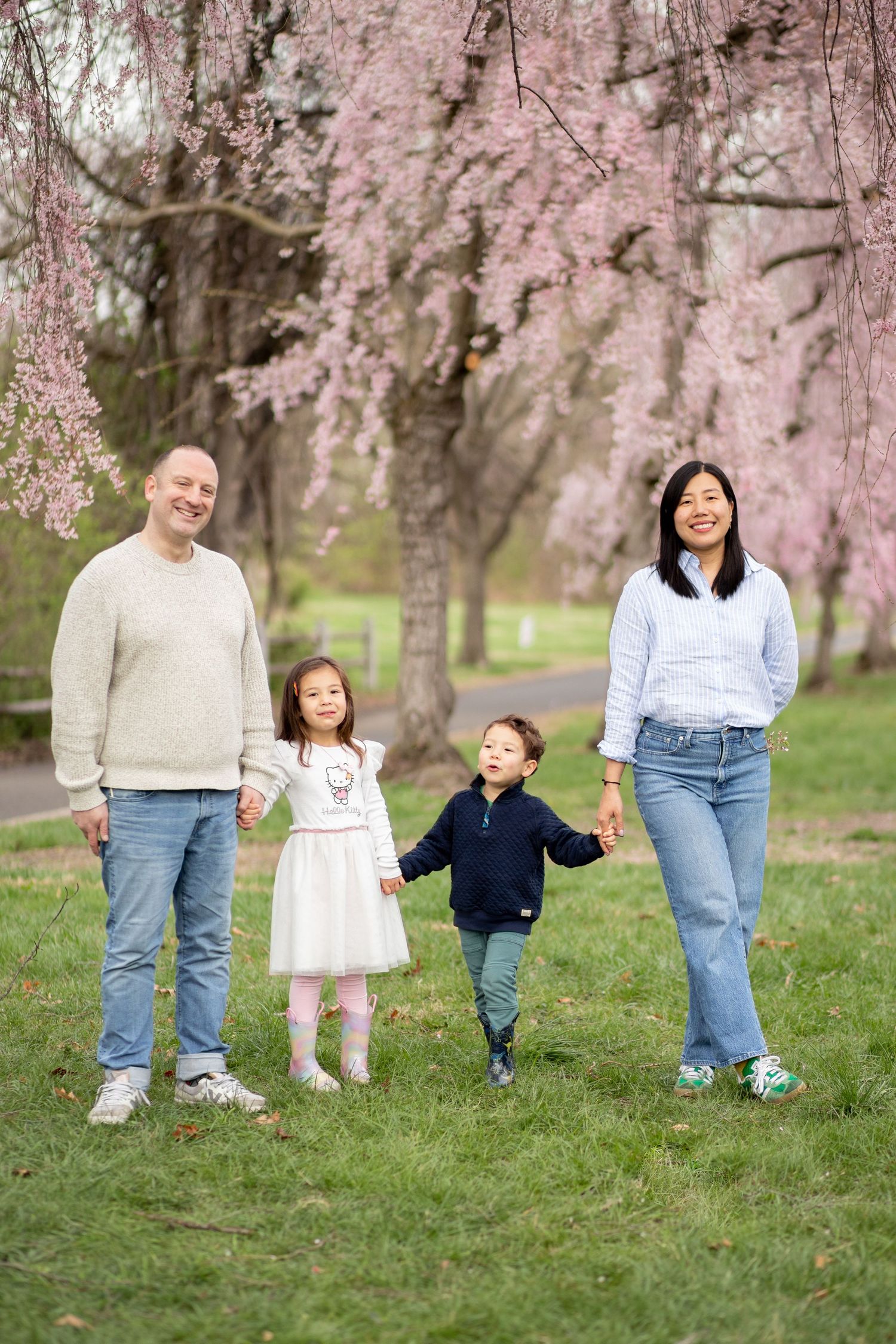 A family stands together on grass beneath blooming pink cherry blossom trees in a spring setting.