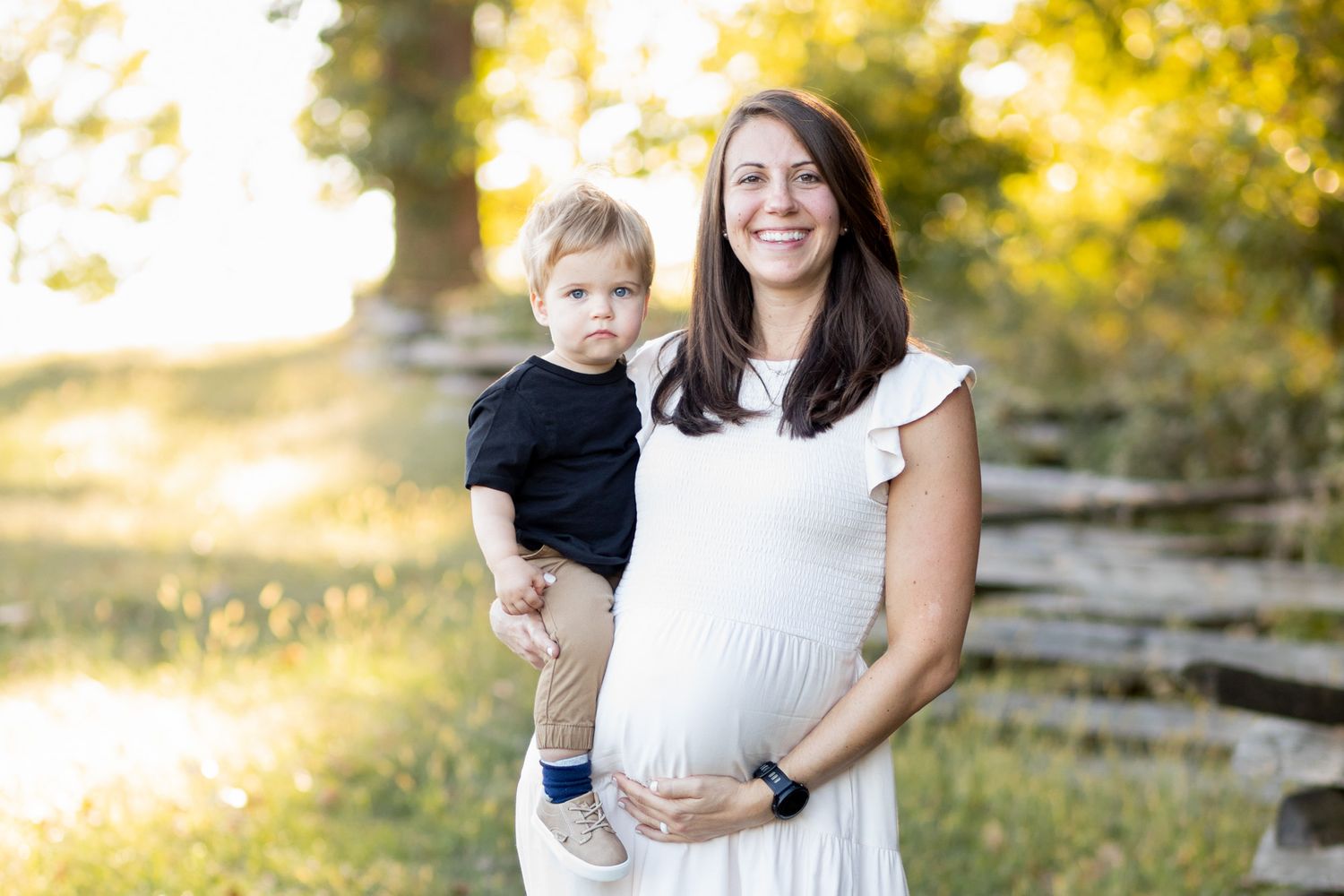 Pregnant woman in white dress holding toddler outdoors in golden sunlight, smiling warmly in a nature setting.