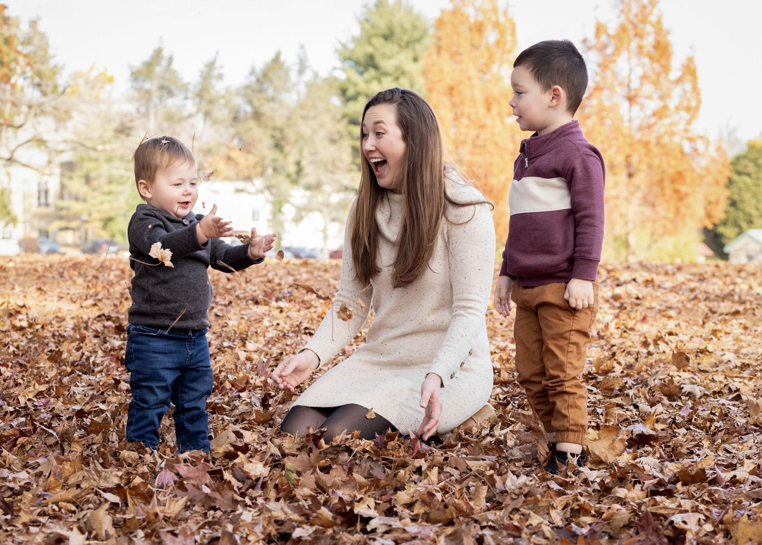 People sitting and playing together in autumn leaves on a sunny day with fall foliage in the background.