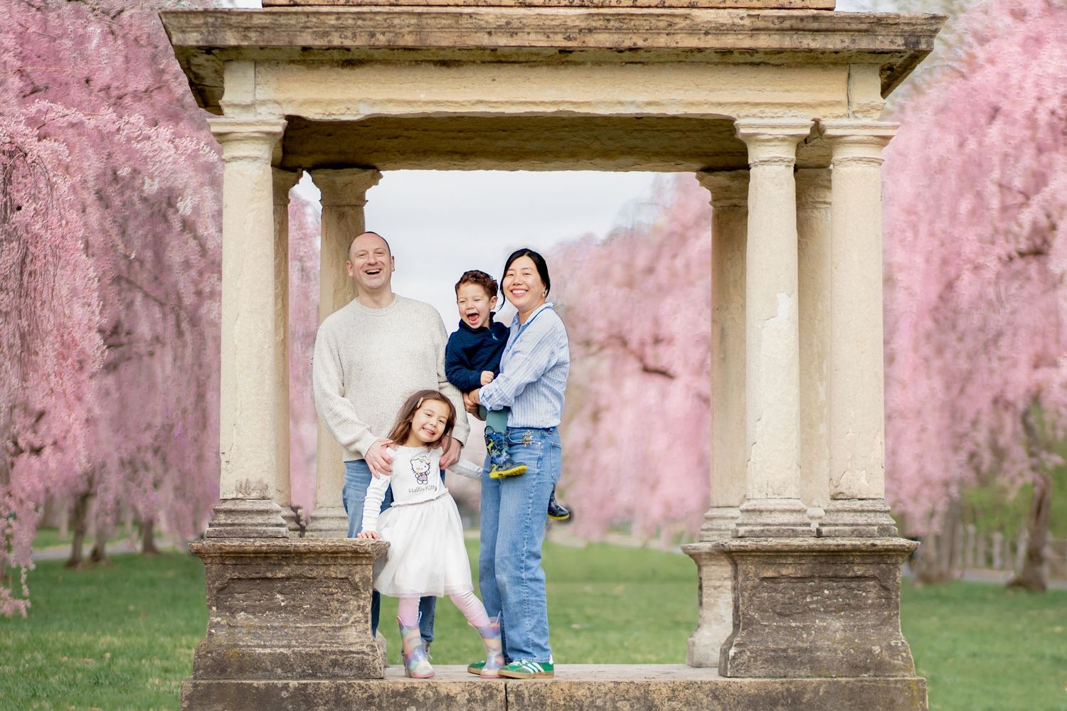 A family stands together under a stone archway with faded pink walls and green grass in a rustic garden setting.