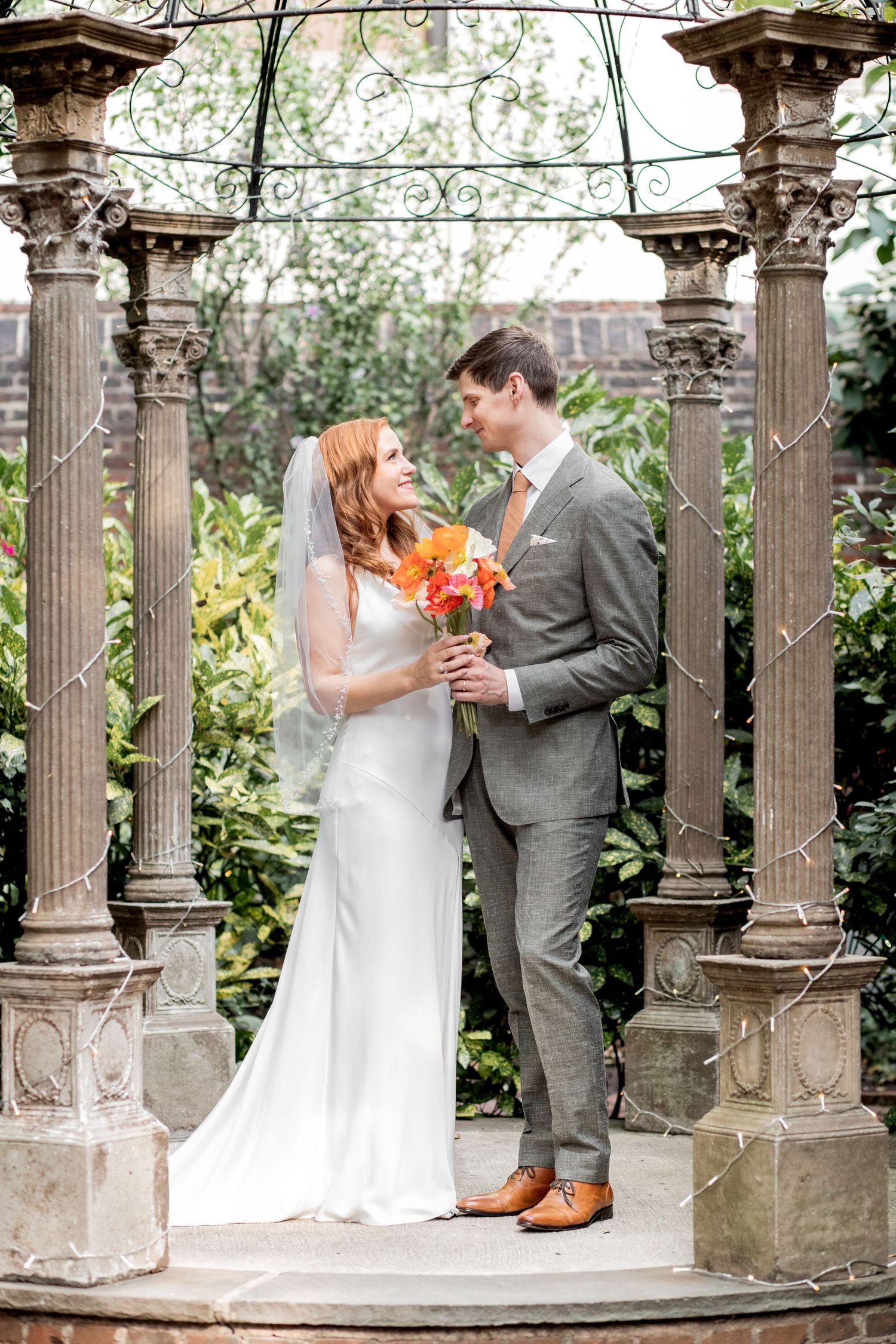 Bride and groom share a romantic moment under ornate garden columns with trailing vines and classic architecture.