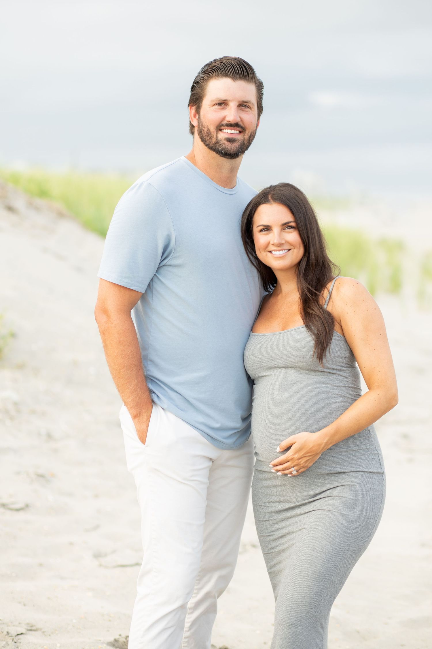 Smiling couple poses on sandy beach, pregnant woman in gray dress with partner in blue shirt and white pants.