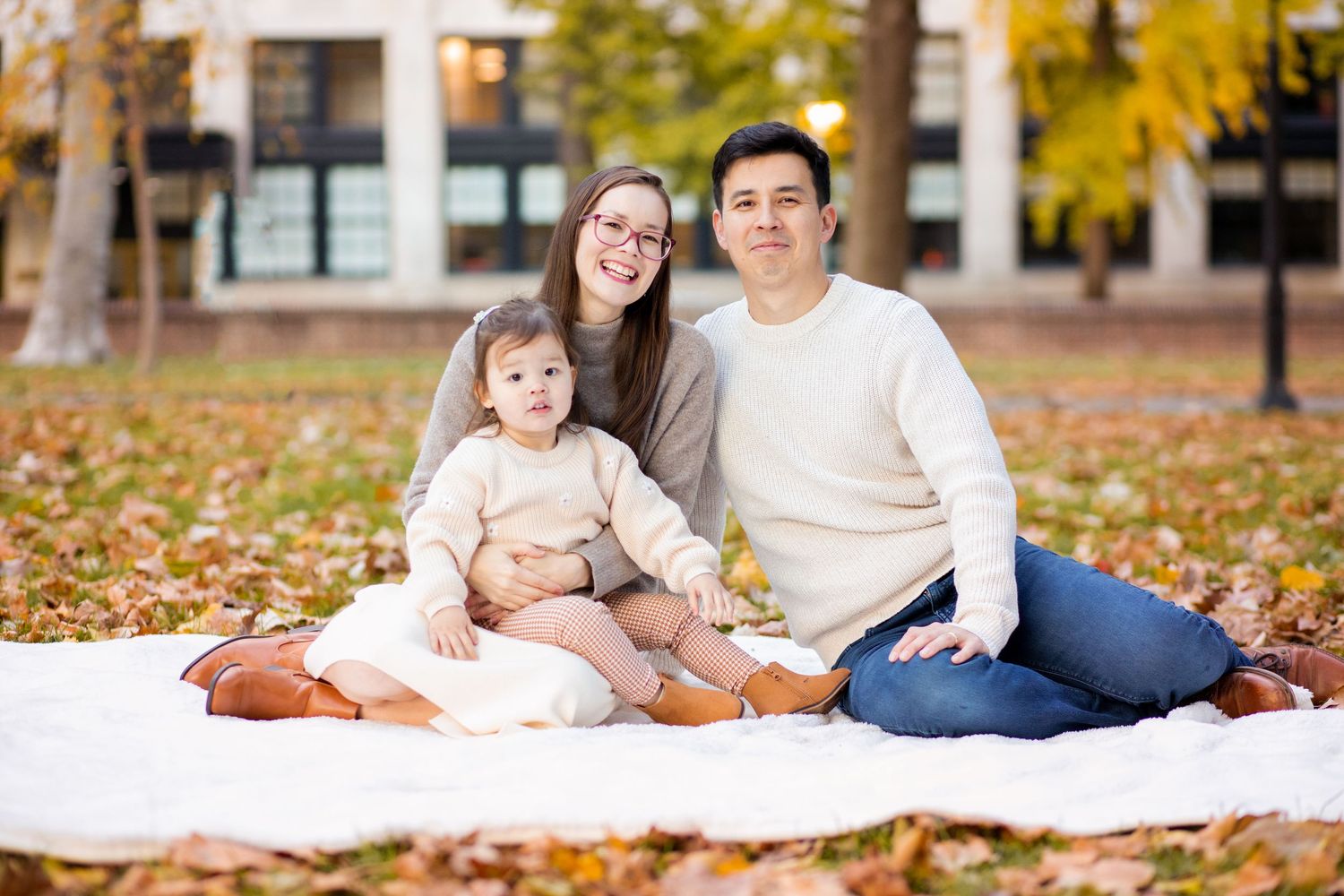 A family sits together on a blanket surrounded by fallen autumn leaves in an outdoor park setting.