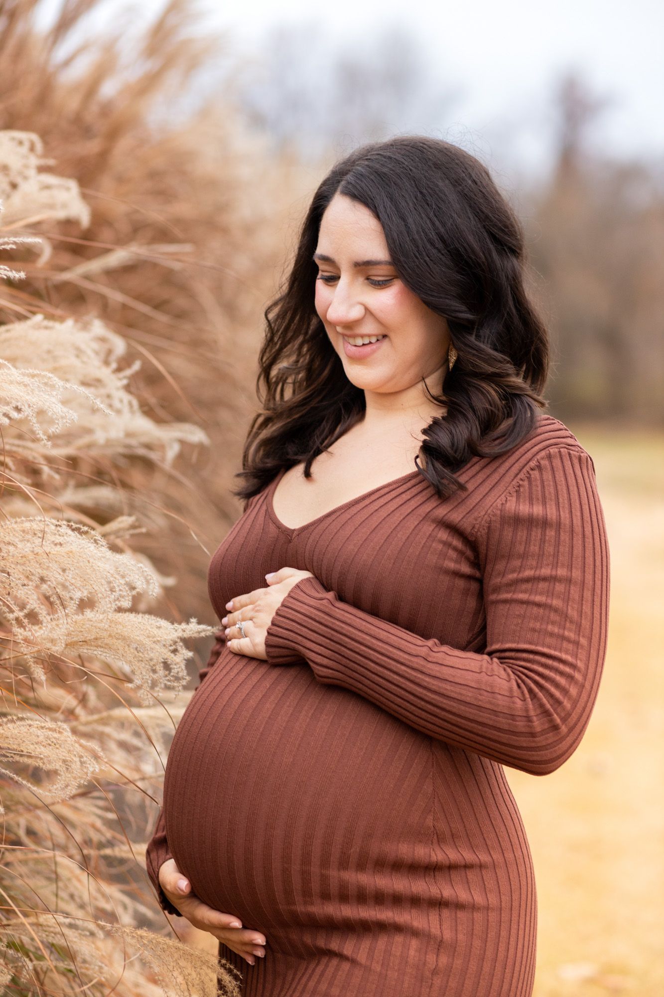 Smiling pregnant woman in brown ribbed dress cradling baby bump near tall golden grasses outdoors.