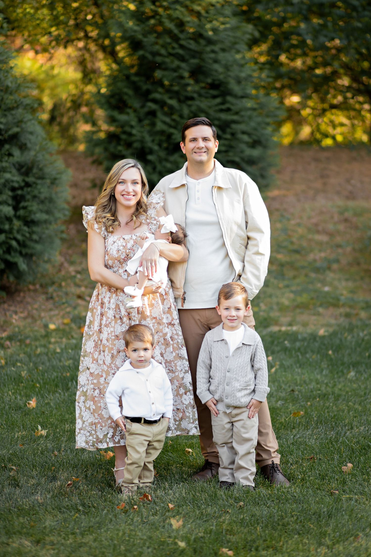 Family of four wearing coordinated neutral tones stands together on grass with trees in background during autumn evening.
