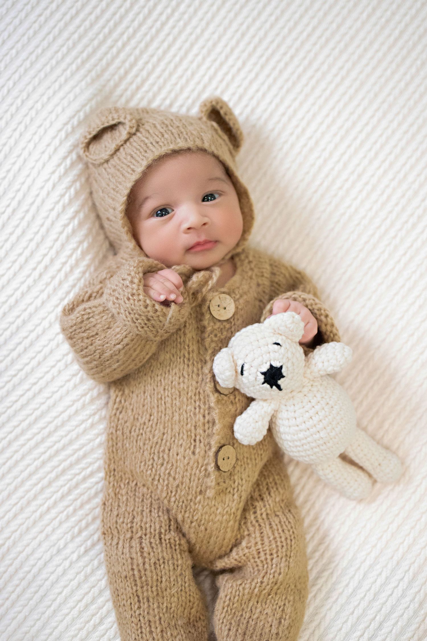 Newborn wearing cozy knitted bear onesie lies on white blanket with matching stuffed polar bear toy.