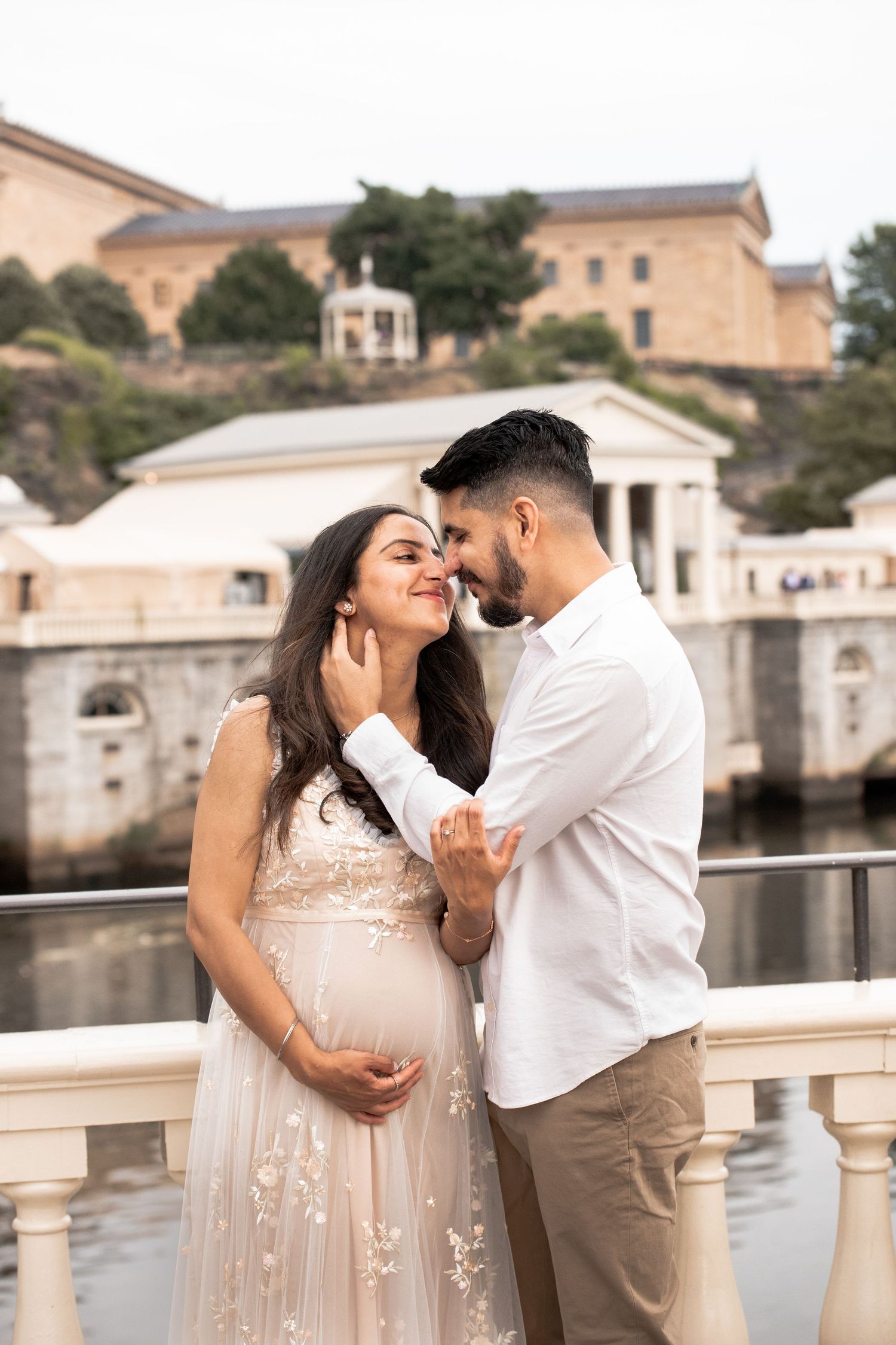 A loving couple embraces by a waterfront balustrade in front of historic Italian architecture, with her wearing a floral dress.