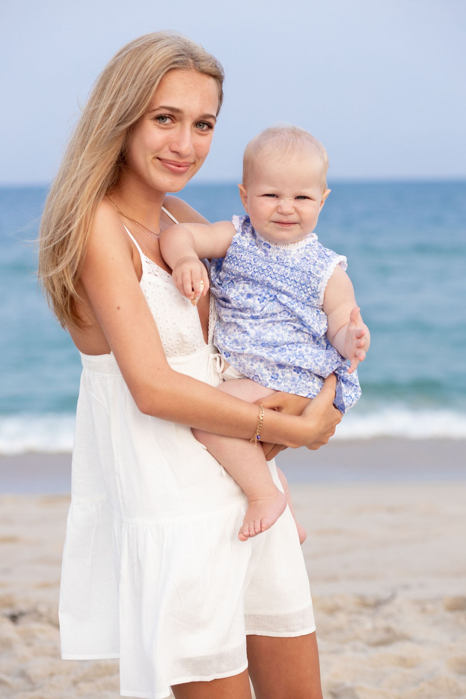 A woman in a white dress holds a baby in a floral outfit on a sunny beach with blue ocean waves in the background.
