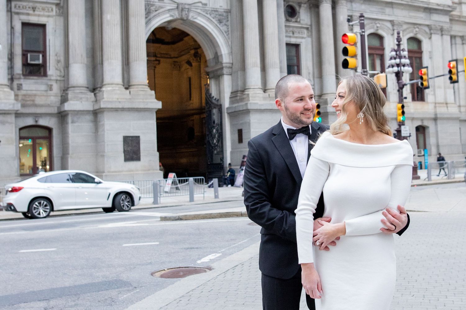 A couple in formal wedding attire stands together on a city street with a grand stone building in the background.