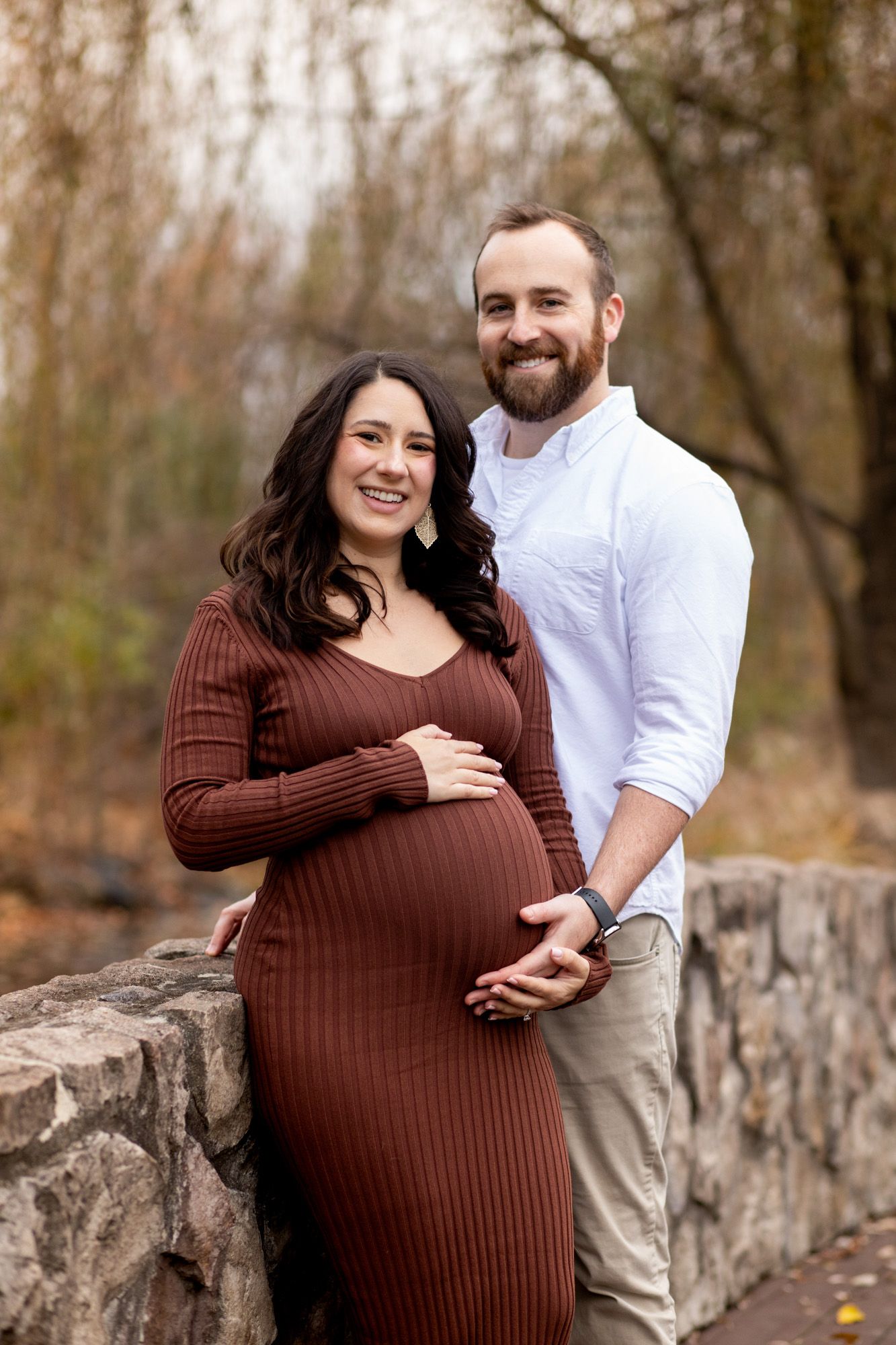 Smiling pregnant couple posing outdoors on a stone bridge, woman in brown ribbed dress, man in white shirt, autumn trees behind.