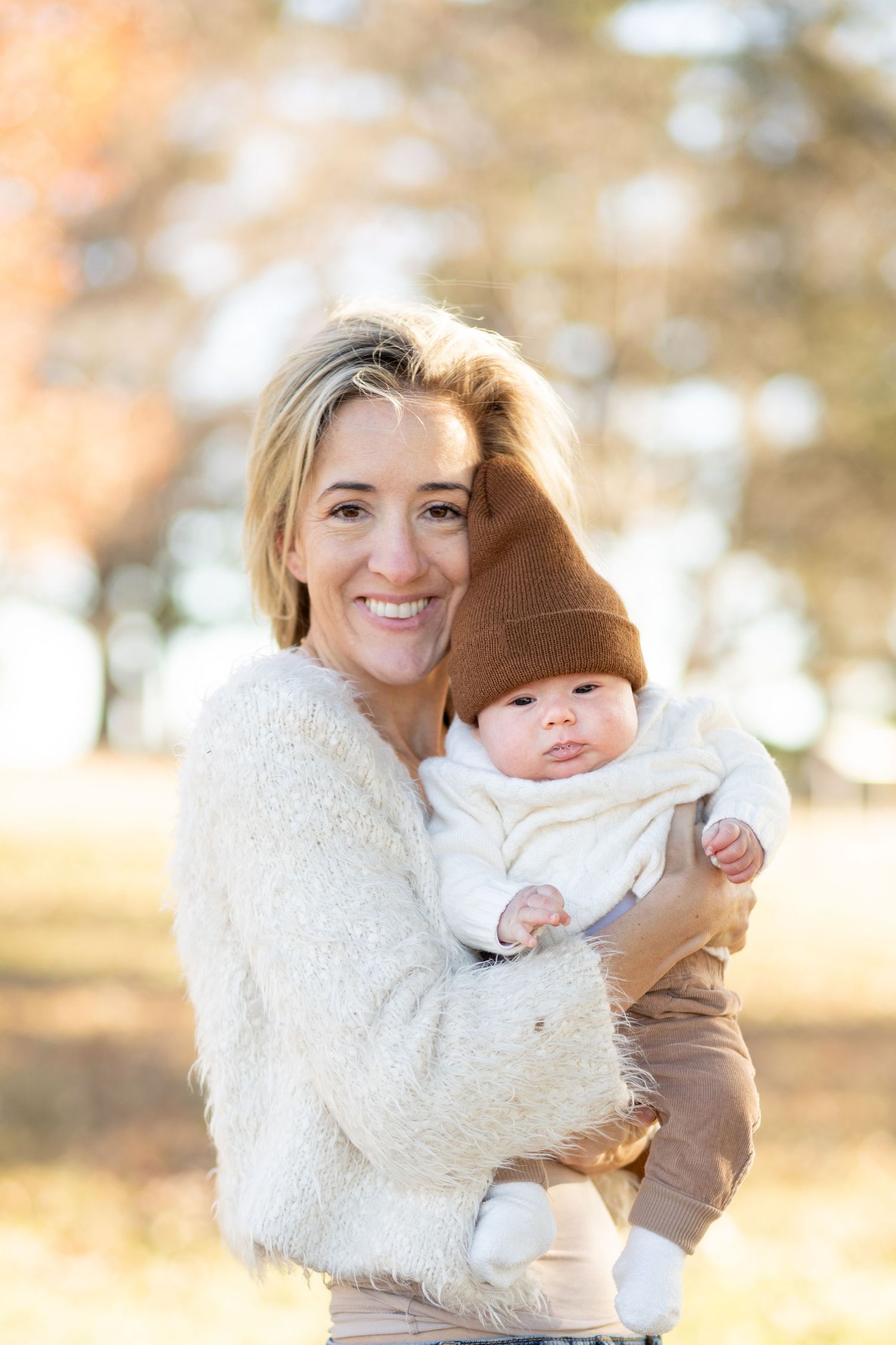 A parent in a white fuzzy sweater holds a baby wearing a brown beanie hat outdoors on a sunny autumn day.