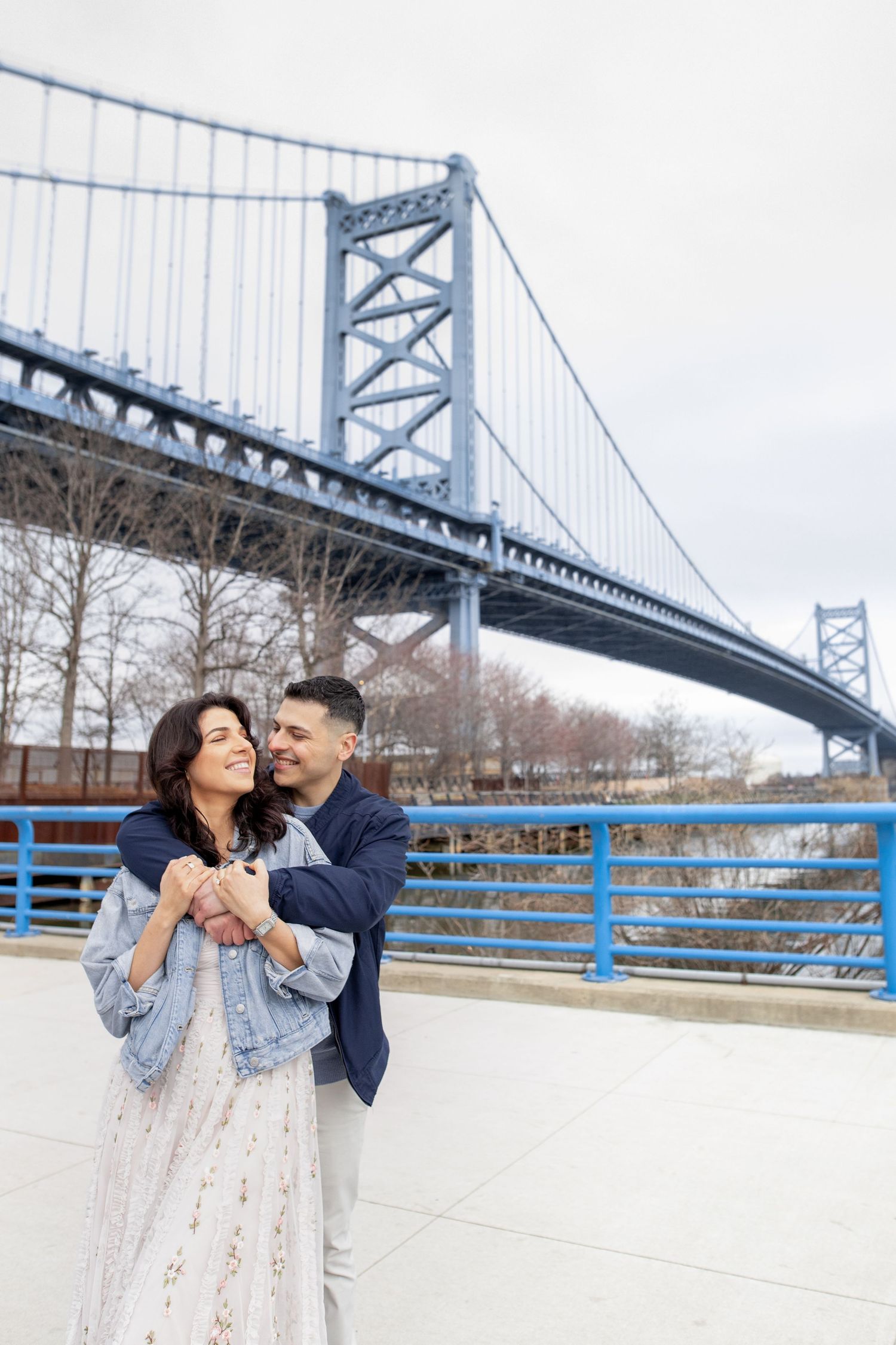 A romantic couple embraces and smiles together under the Benjamin Franklin Bridge in Philadelphia during winter.