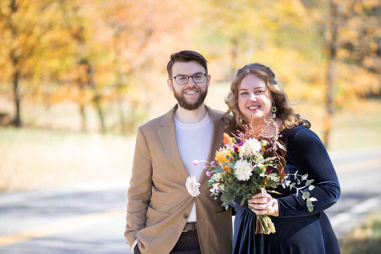 A happy couple stands outdoors during autumn, with one person holding a colorful bouquet against a golden foliage backdrop.