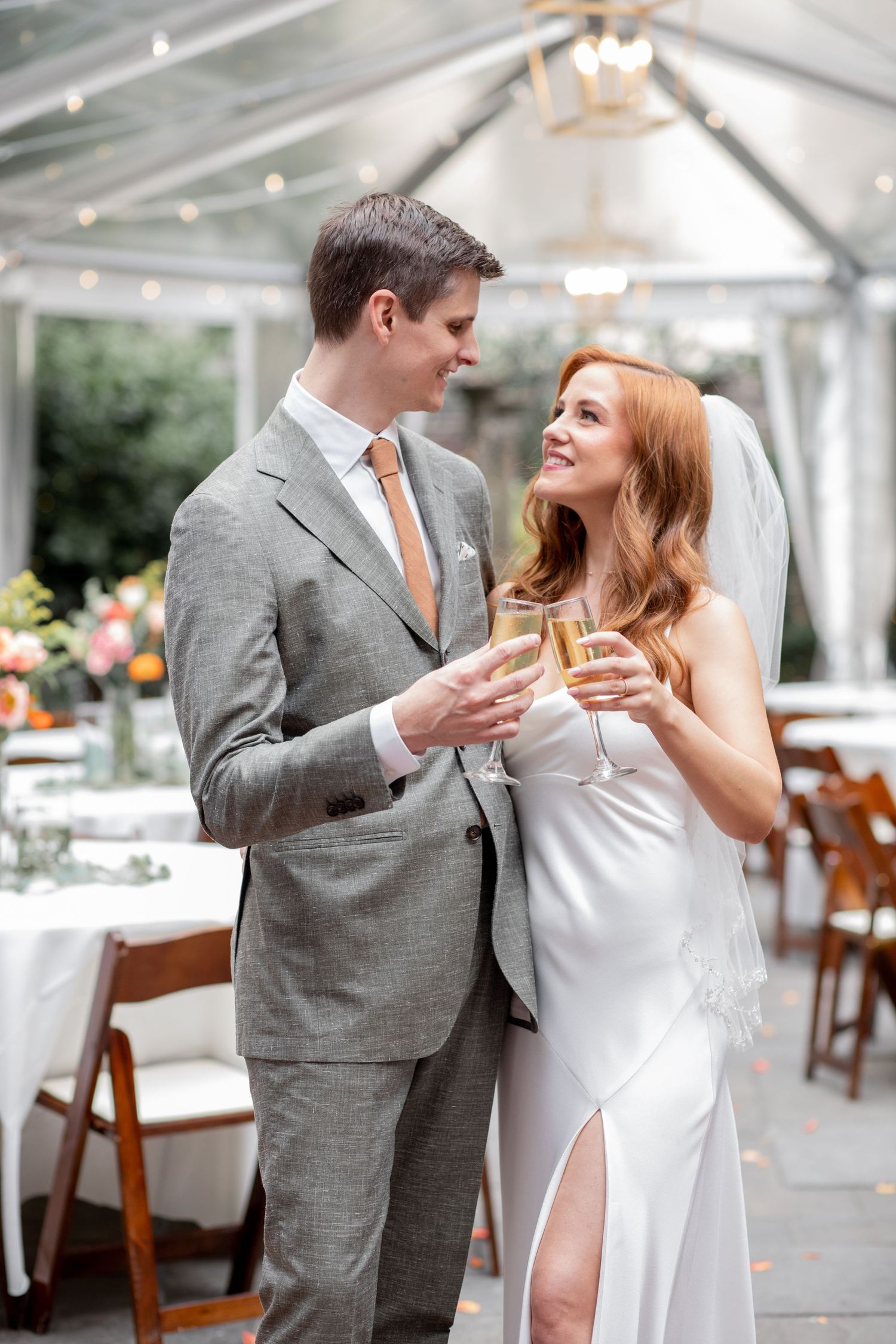 A couple in formal attire share a romantic moment at an elegant wedding reception with a white tent in the background.