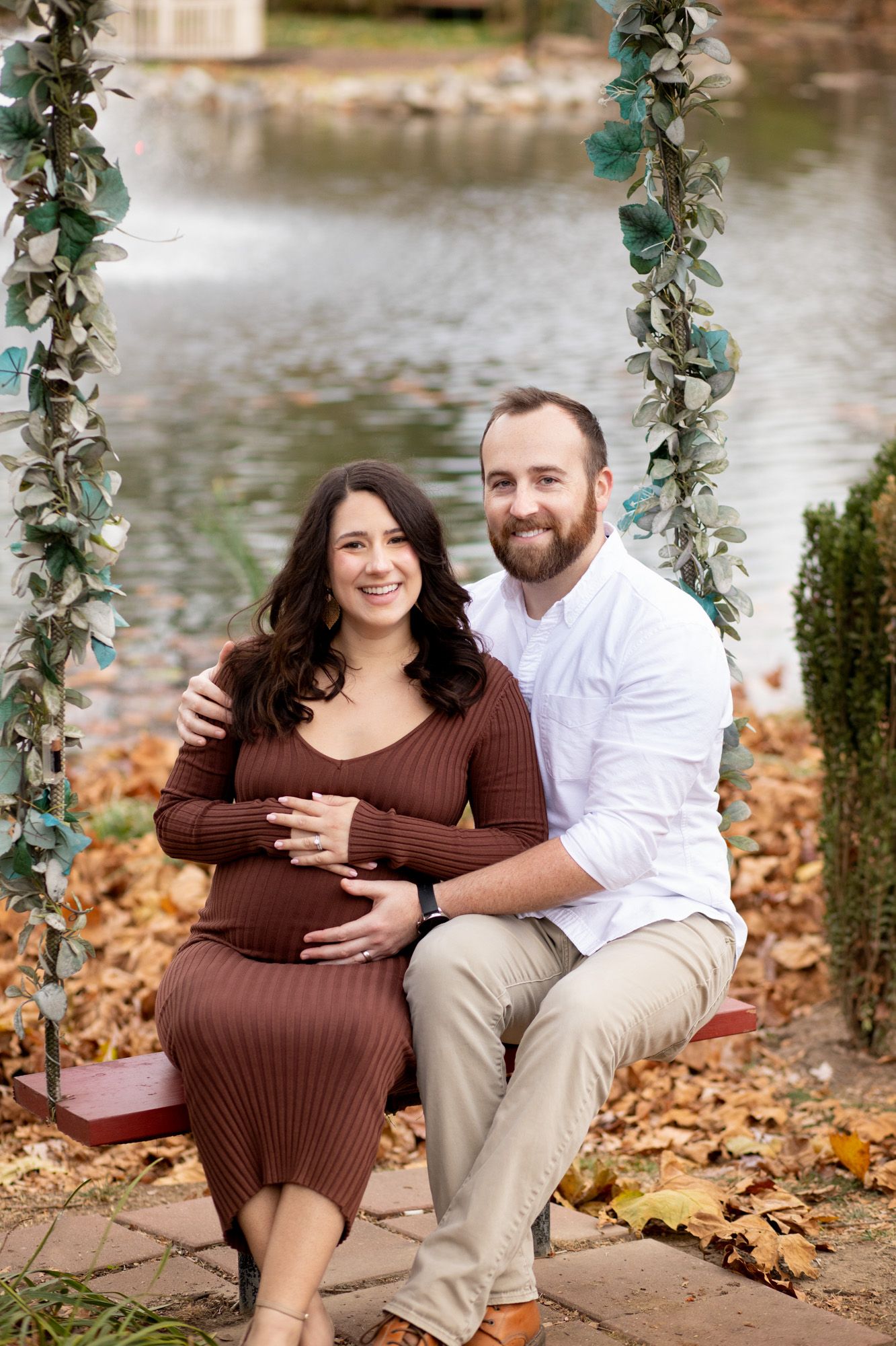 Smiling pregnant couple sitting on a swing decorated with greenery near a pond during autumn.