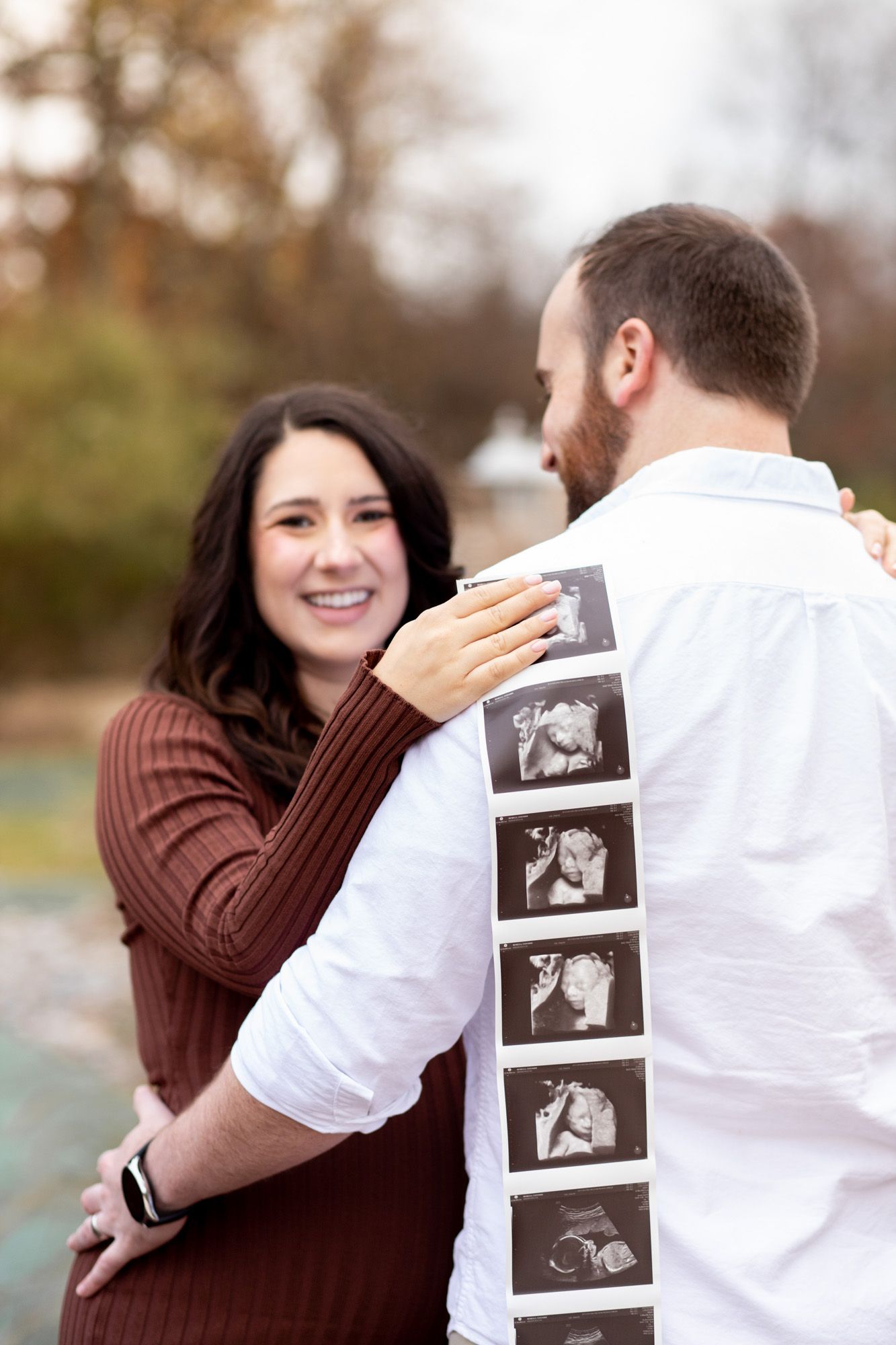 Couple sharing a joyful pregnancy announcement moment outdoors, man holding ultrasound photos on his back.