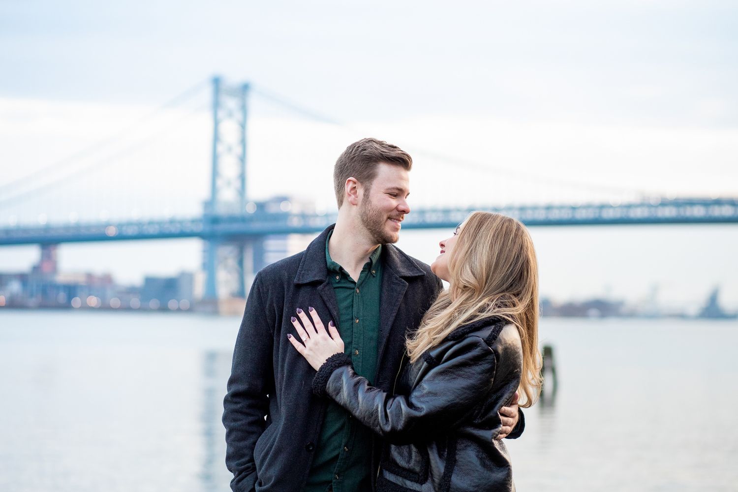 A couple embraces and smiles at each other by the waterfront with the Benjamin Franklin Bridge in Philadelphia visible behind them.