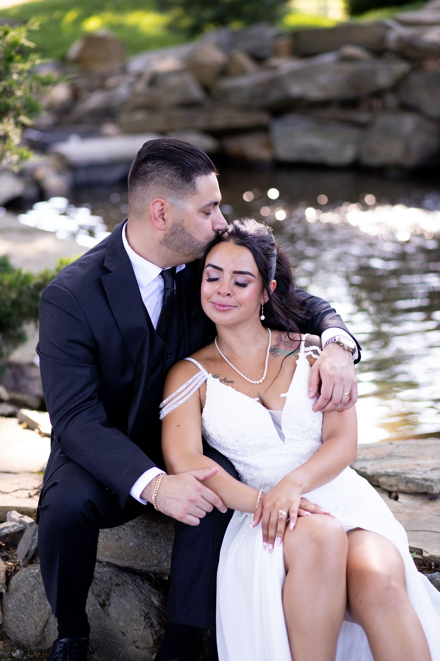Couple in formalwear share intimate moments by a waterside rock garden in romantic wedding photo series.